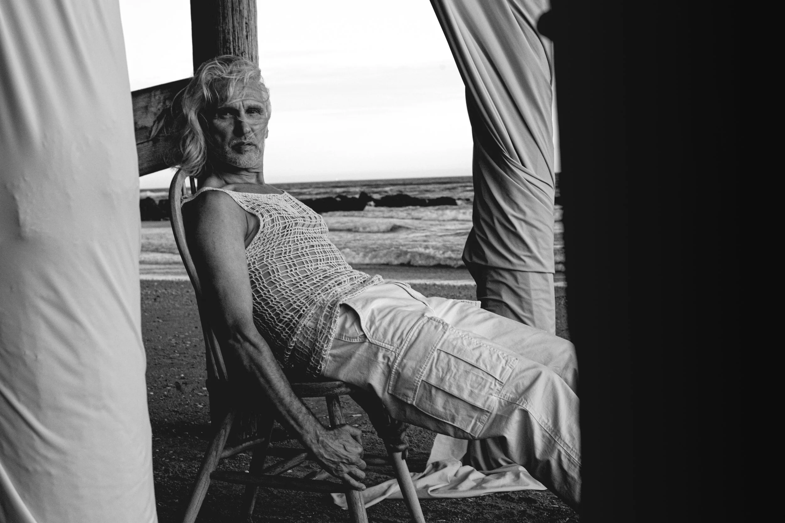 A black and white photo of an older man relaxing on a beach, sitting in a chair with a sundress and shorts. The man is reclining and looking toward the camera, with the ocean in the background and a beach at sunset or sunrise.