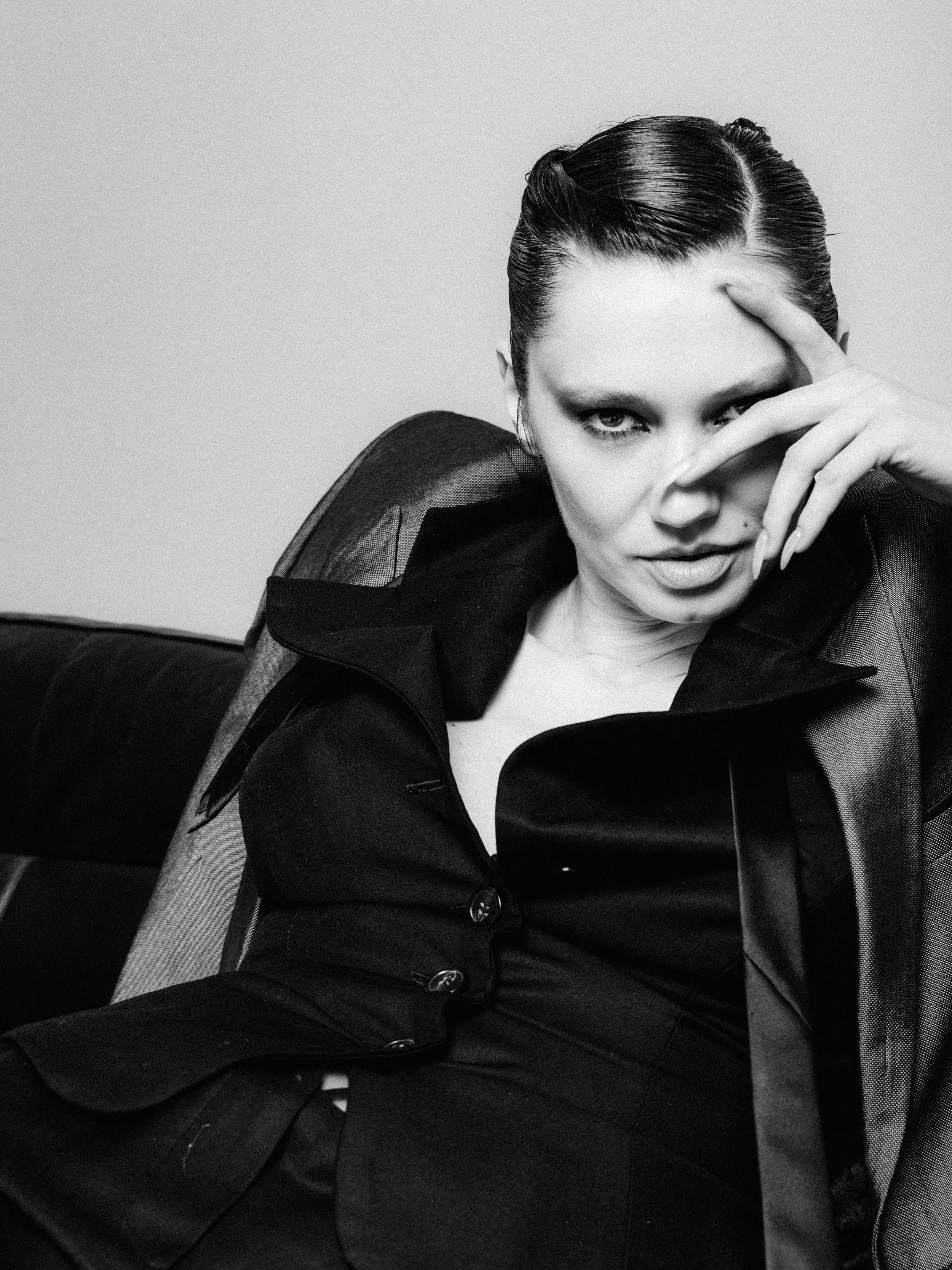 A black and white photo of a woman with short, slicked-back hair, wearing a dark blazer with sheer shoulder details, sitting on a black leather surface, looking directly at the camera with one hand partially covering her face.