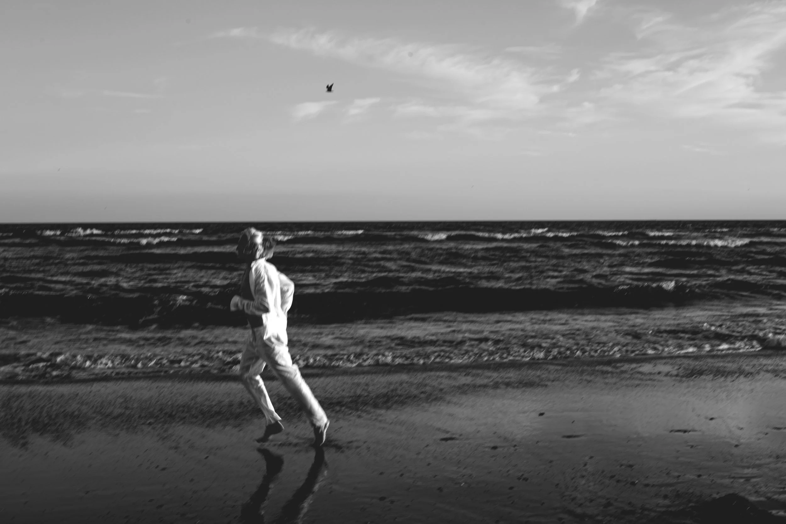 A young boy running along the beach, wearing light-colored clothes, with the ocean waves in the background and a bird flying in the sky.