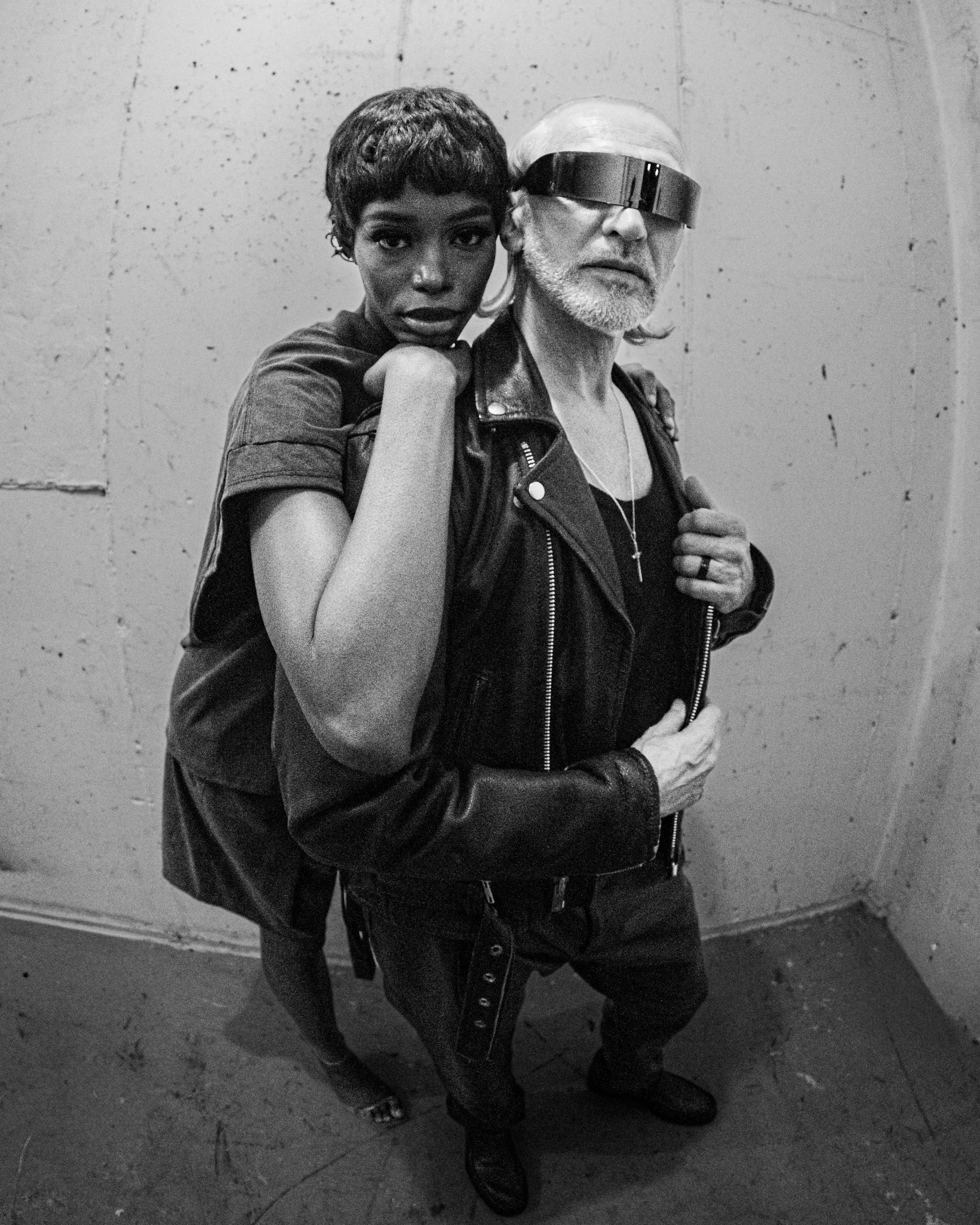 A black and white photo of a woman and an older man with special eyewear, standing close against a concrete wall.