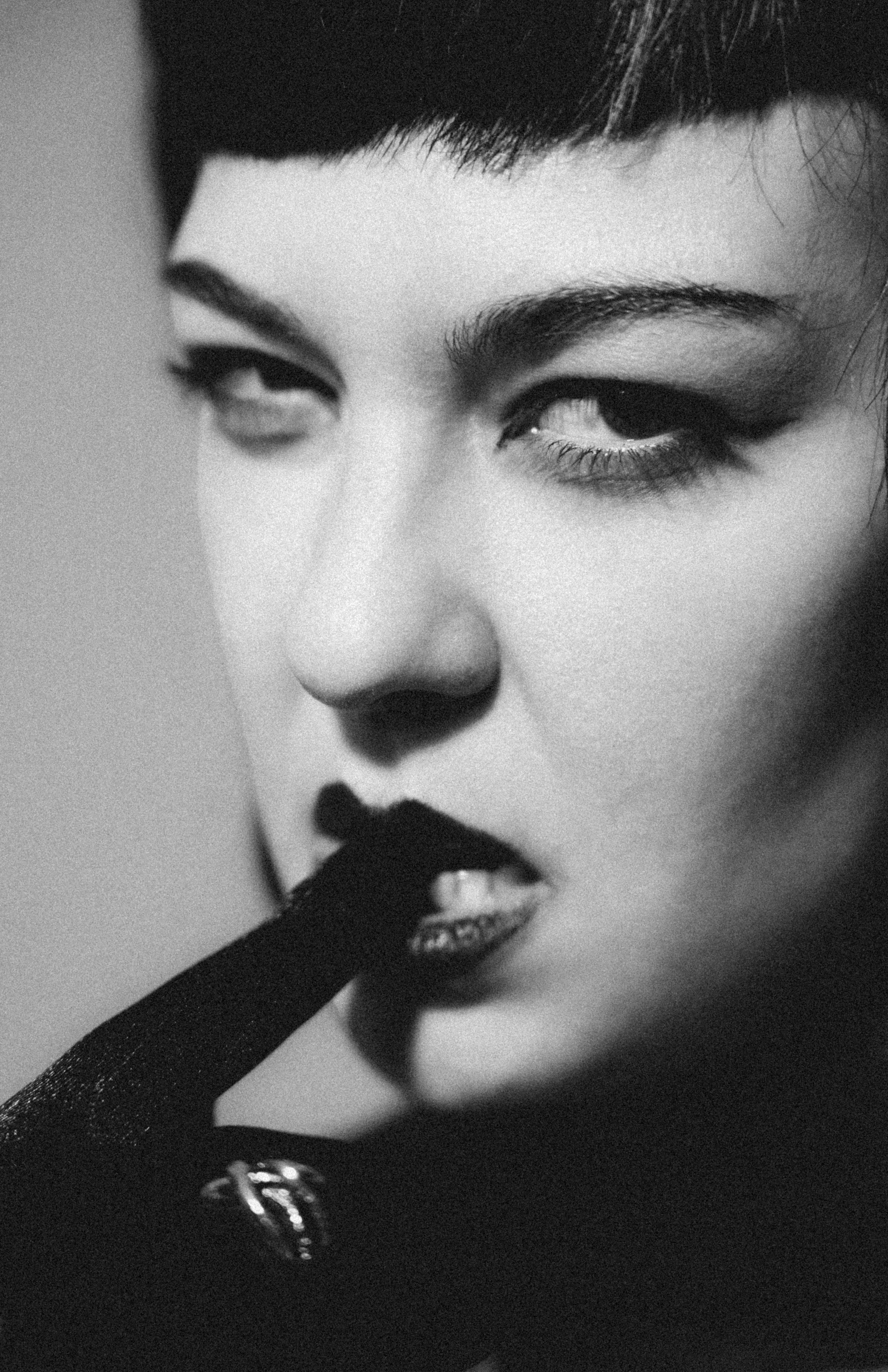 Close-up black-and-white photograph of a woman's face with dark eye makeup and lipstick, wearing a satin glove and a ring, with her finger touching her lips.
