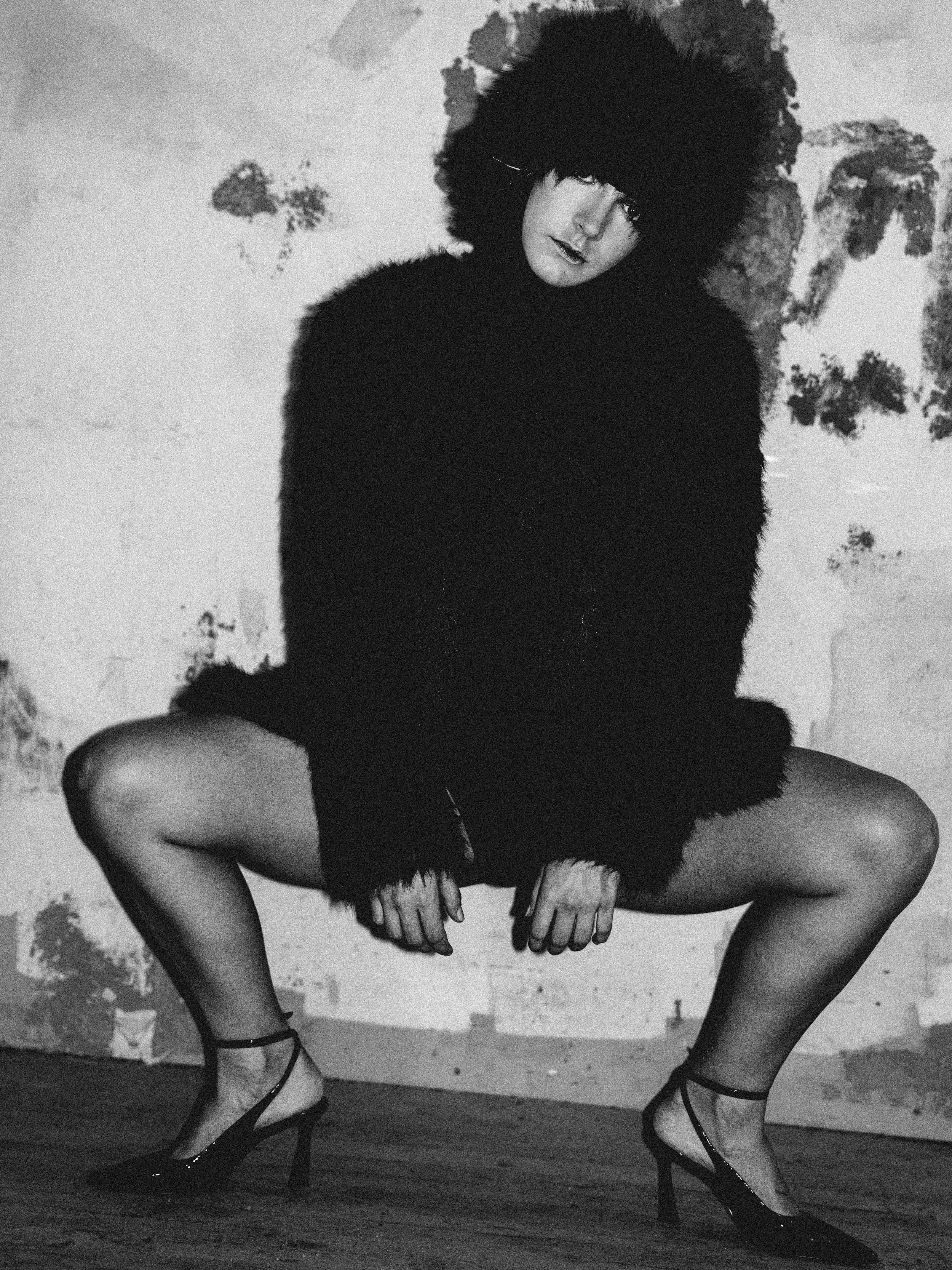 A woman in high heels, sitting with her legs apart, wearing a black fuzzy coat with a large hood that covers her head and face, against a distressed wall background.
