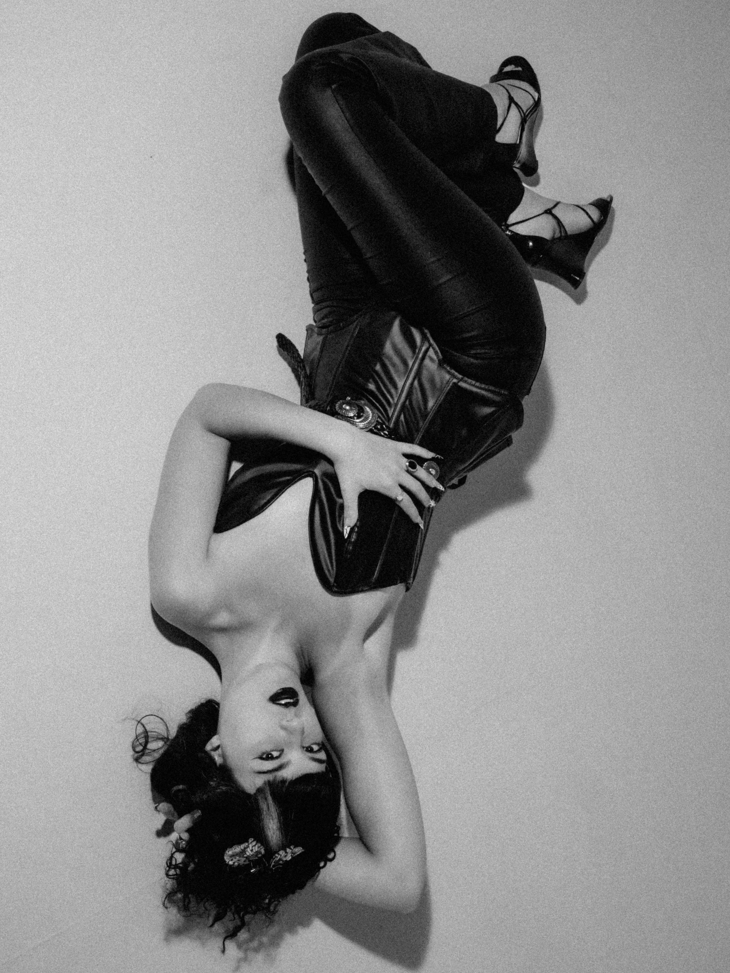 A black and white photo of two women in opposite poses. One woman with curly hair is lying on her side, facing the camera, wearing a dark lace outfit. The other woman, with straight hair, is sitting with her back to the camera and her head turned to 