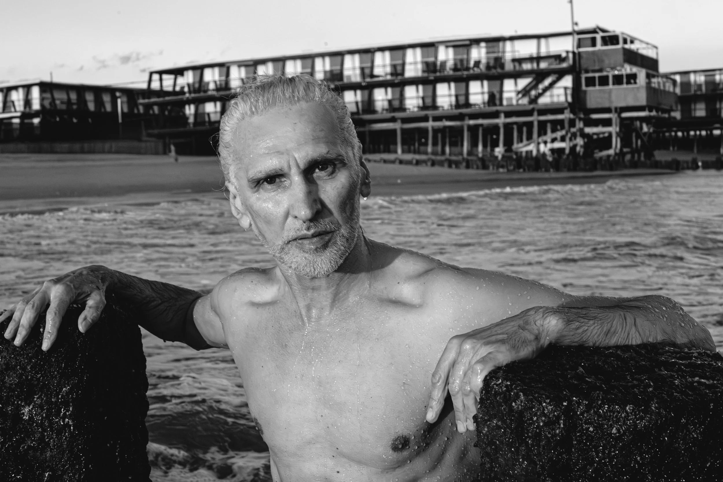 A man with wet hair and a beard, partially submerged in water, appears to be emerging from the ocean or a large body of water with a pier or boardwalk in the background.