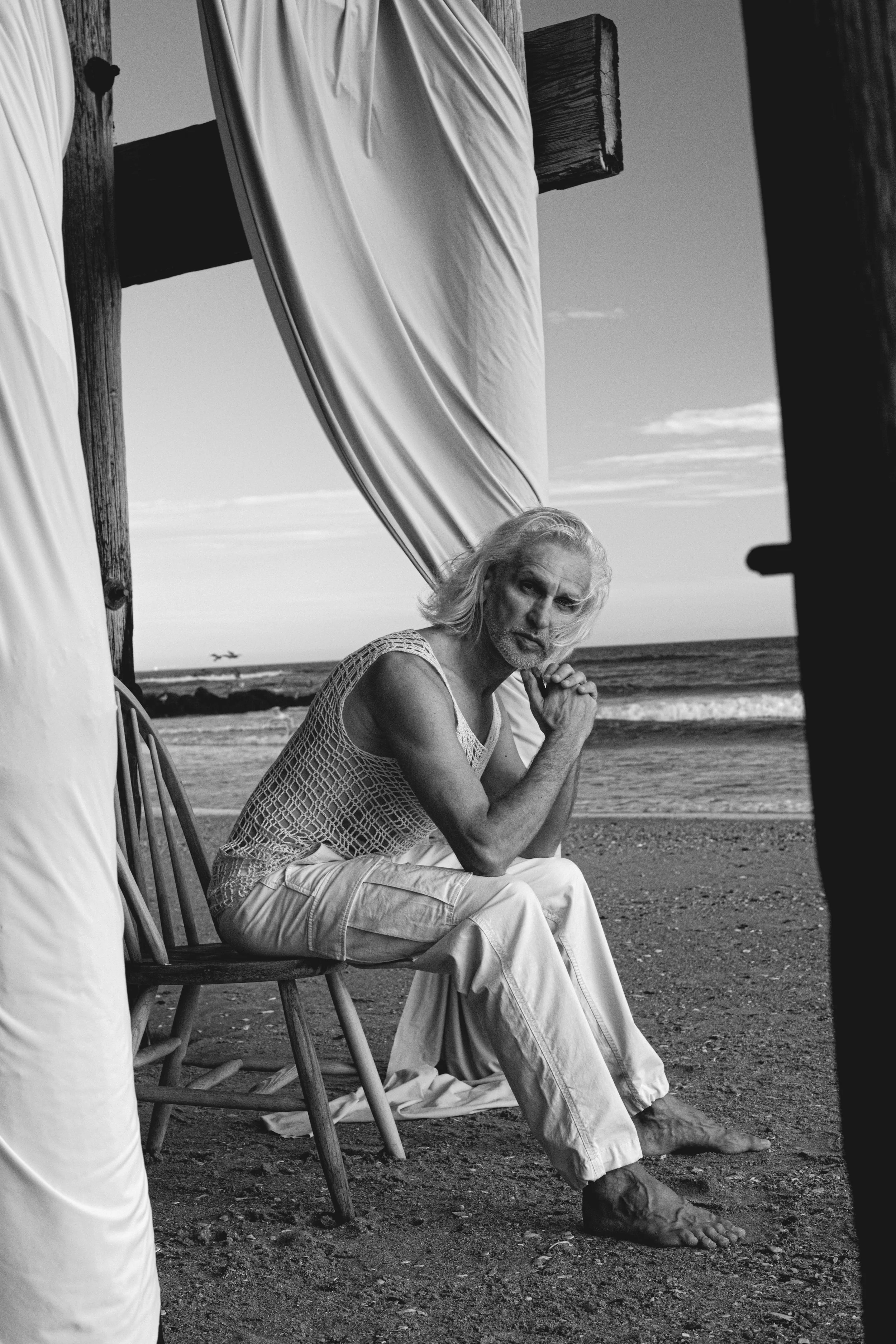 A black and white photo of an elderly man with long hair and a beard, sitting on a wooden chair on a beach. He has a pensive expression and is dressed in a sleeveless mesh shirt and light-colored pants. The scene includes a large umbrella and the oce