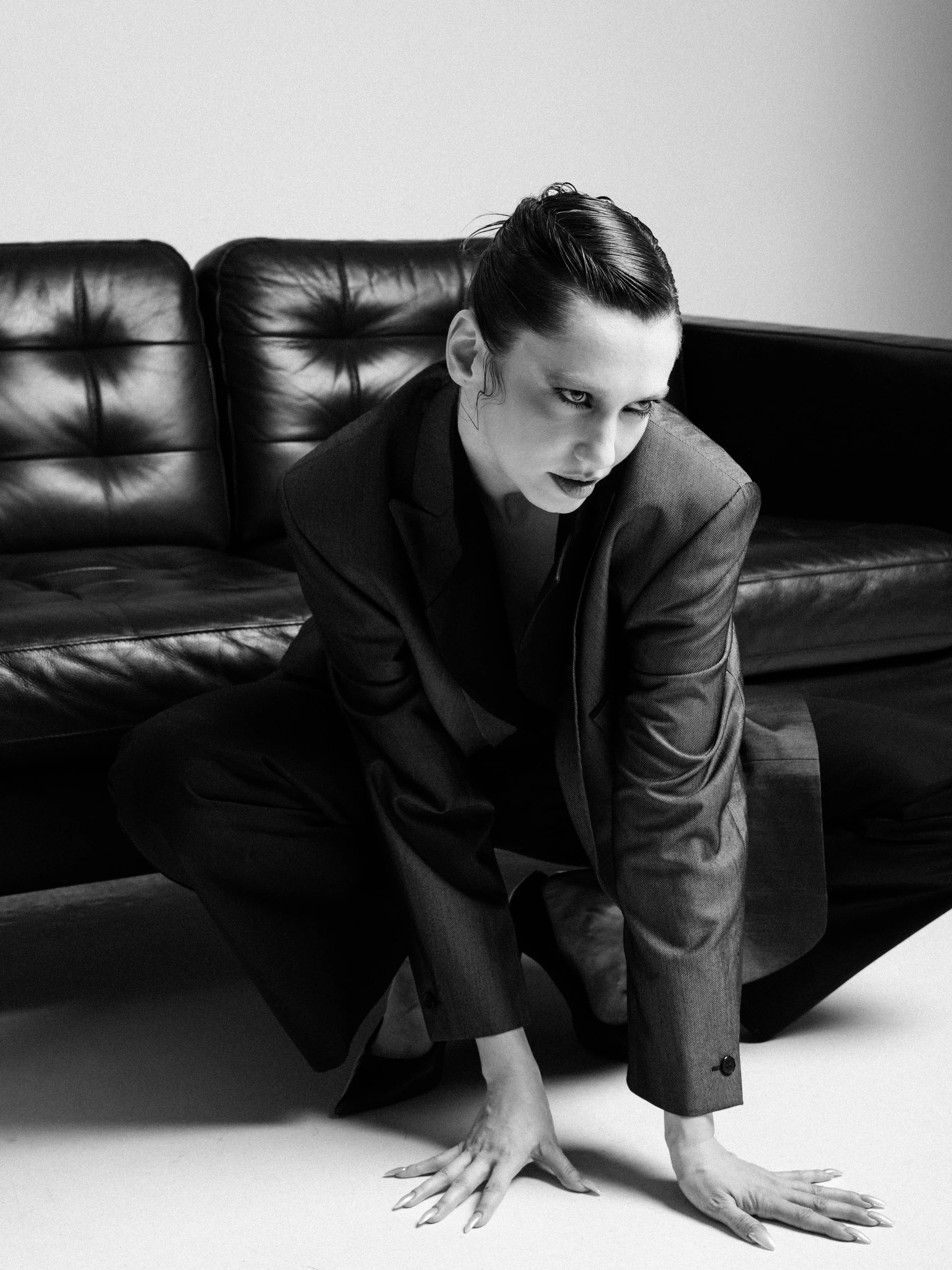 Black and white photo of a woman with dark, slicked-back hair wearing a suit, posed on the floor with her hands flat and fingers spread out, near a leather couch against a plain wall.