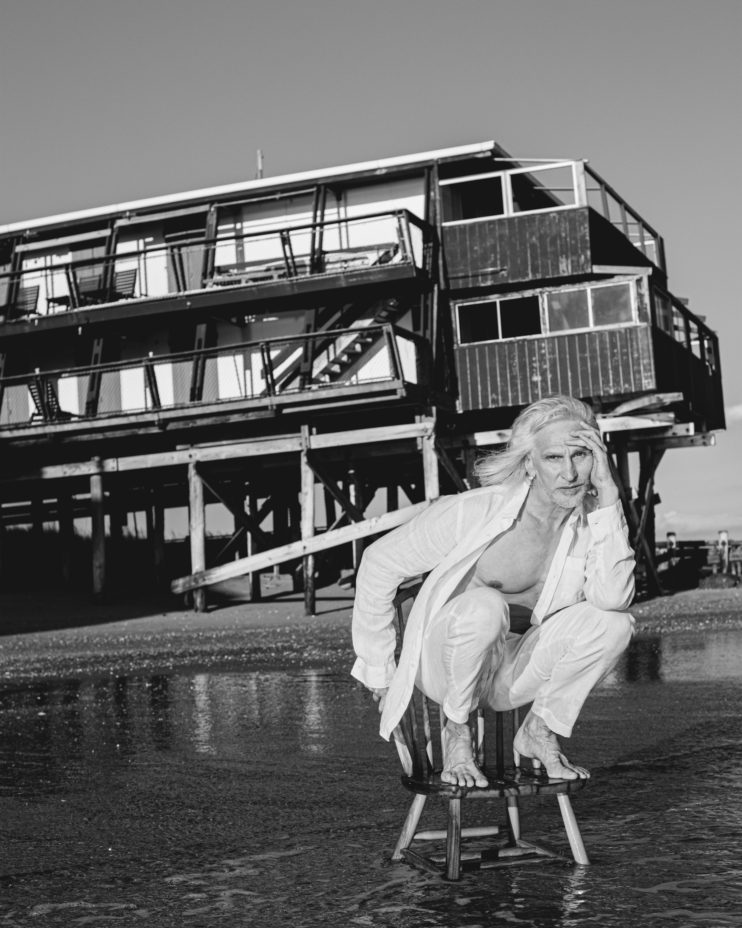 A woman with long hair and a white shirt crouching on a chair with her hand on her head, near a shoreline with a large wooden structure and an apartment building in the background.