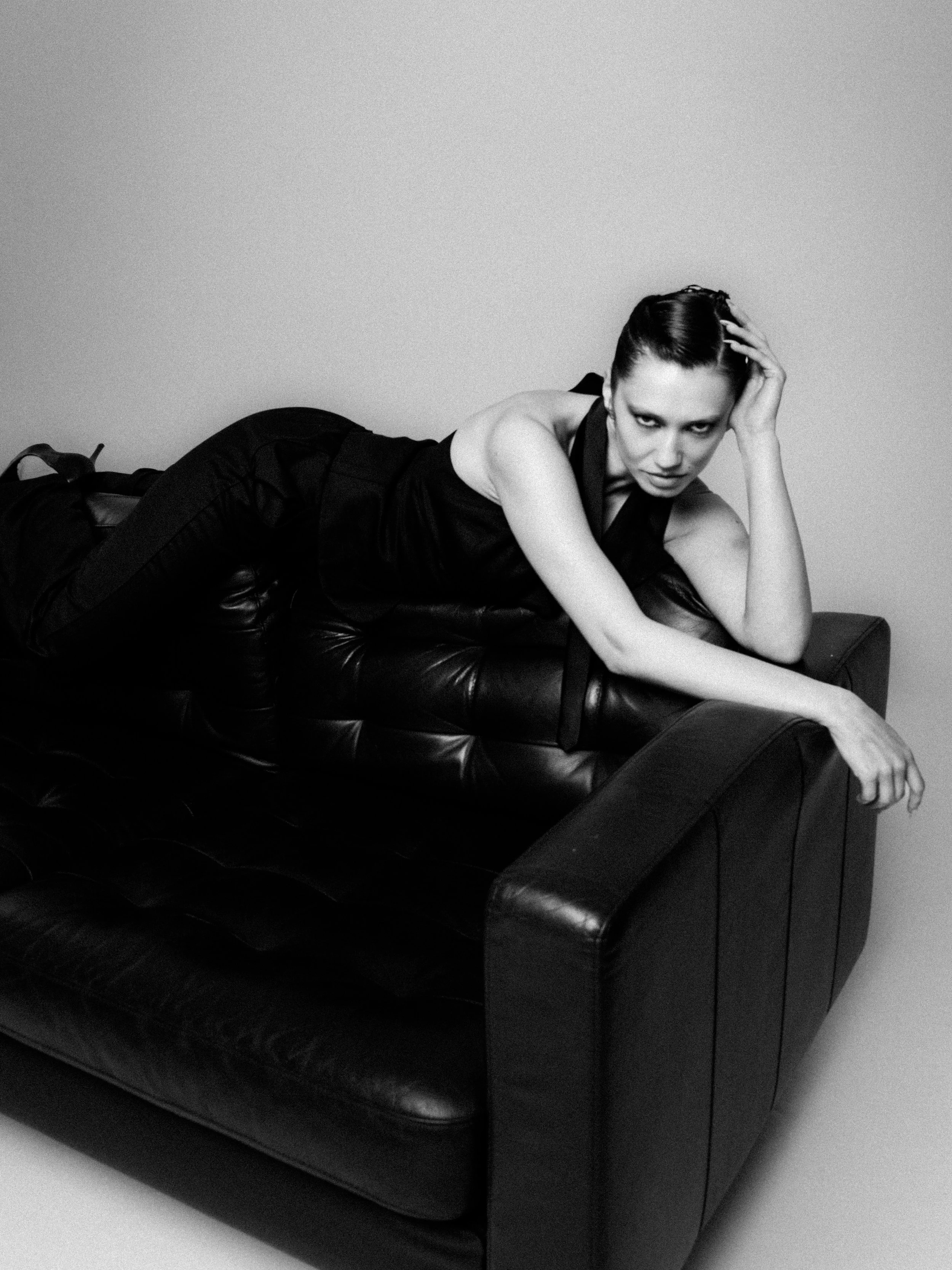 Black and white photo of a woman lying on a leather couch, resting her head on her left hand, looking directly at the camera with a serious expression.