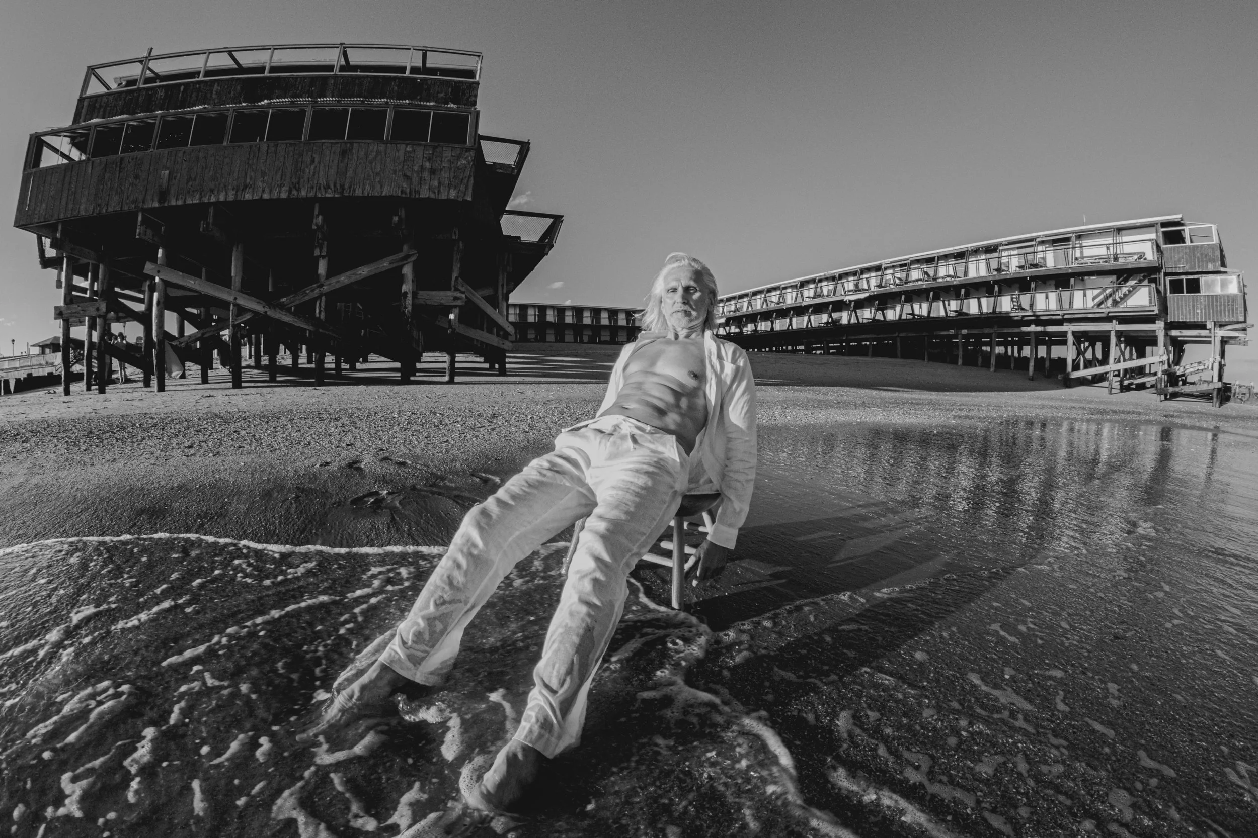 A man with long hair and a beard, dressed in white, sitting on a beach near the water with waves reaching his legs. Behind him, there are large, elevated wooden structures or boardwalks on stilts, and the sky is clear.