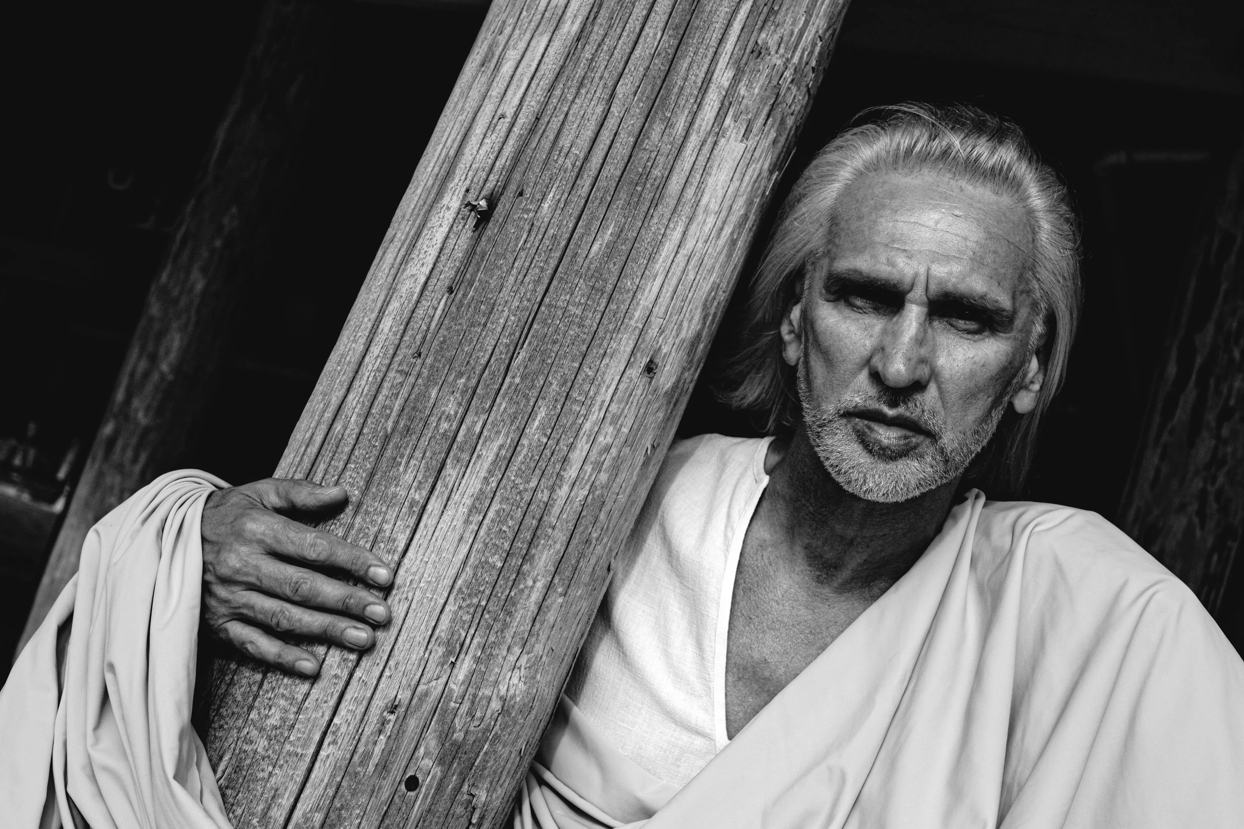 Black and white photo of a man with gray hair and a beard holding a wooden beam on his shoulder.