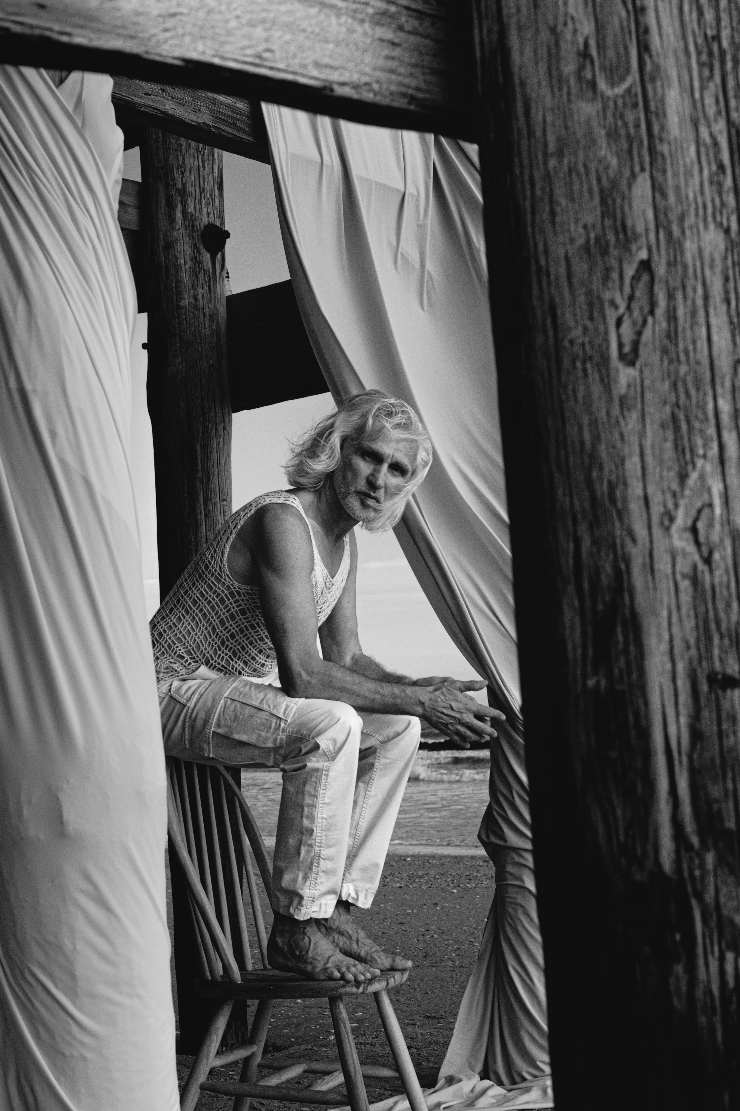 A black and white photo of an older man with long gray hair, sitting on a wooden chair outdoors, next to a tree, capturing a contemplative moment as he rests his hand on his knee. The man is framed by wooden beams and light-colored drapery.
