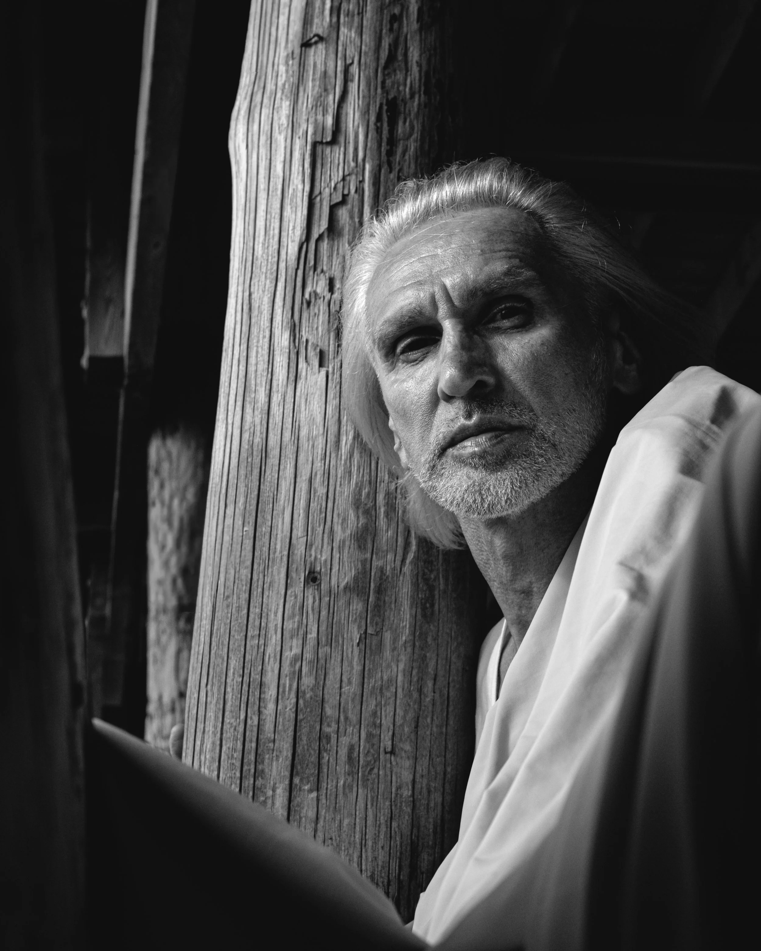 Black and white photo of an elderly man with white hair and a beard, lying down next to a weathered wooden post, with his hand resting on his head.