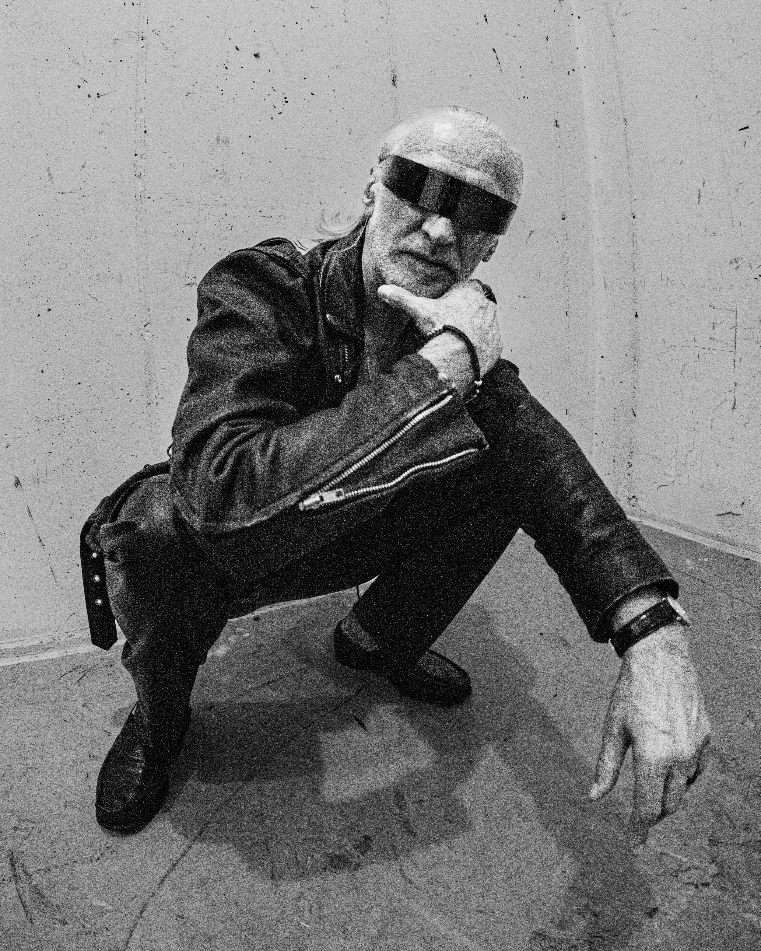 Black and white photo of an older man with long hair and beard wearing sunglasses, leather jacket, and boots, crouching against a wall with his right hand resting on his chin.