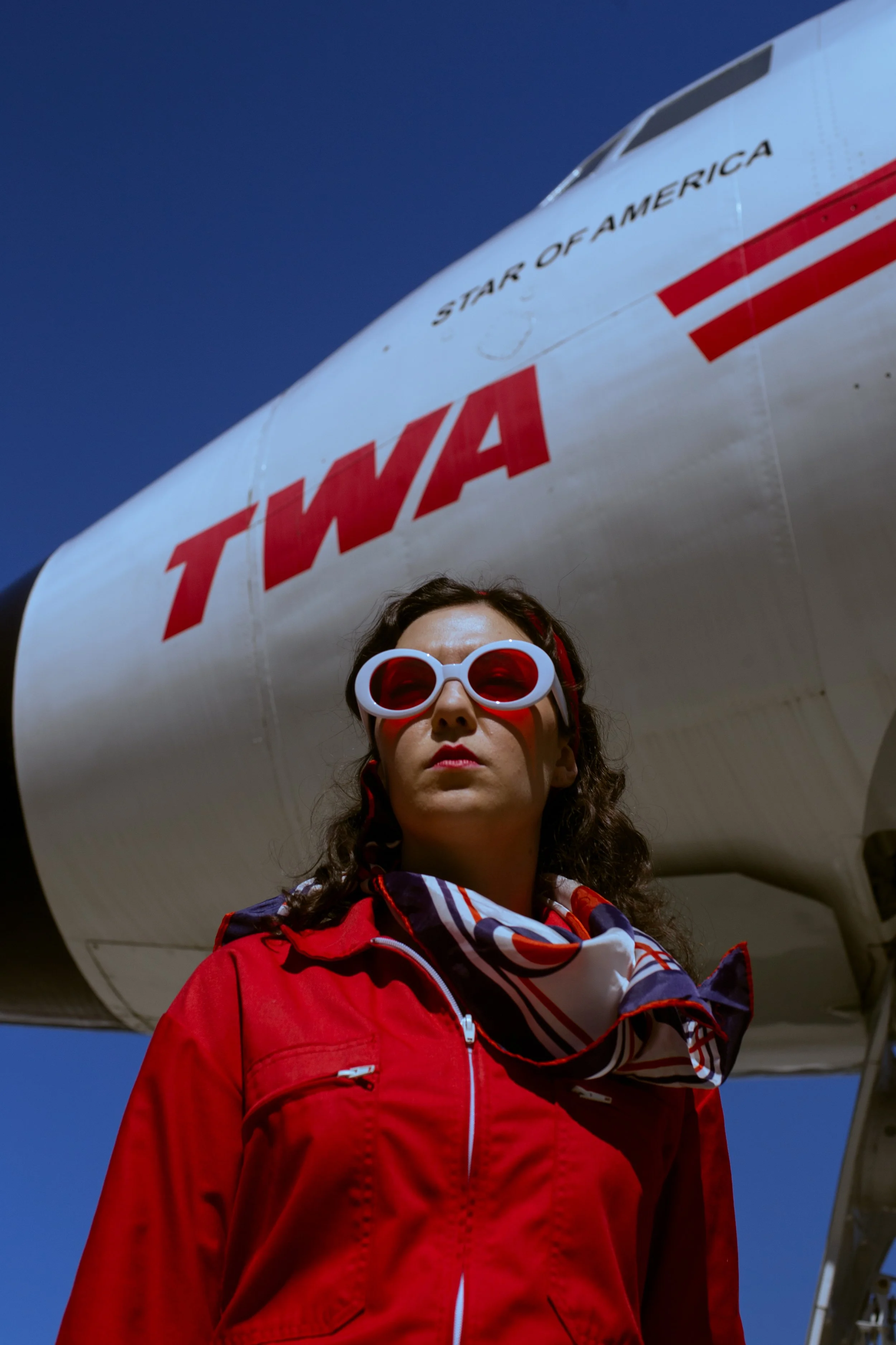 A woman with curly hair wearing sunglasses, a red jacket, and a scarf, standing in front of an airplane with the words 'Star of America' and 'TWA' on its fuselage.