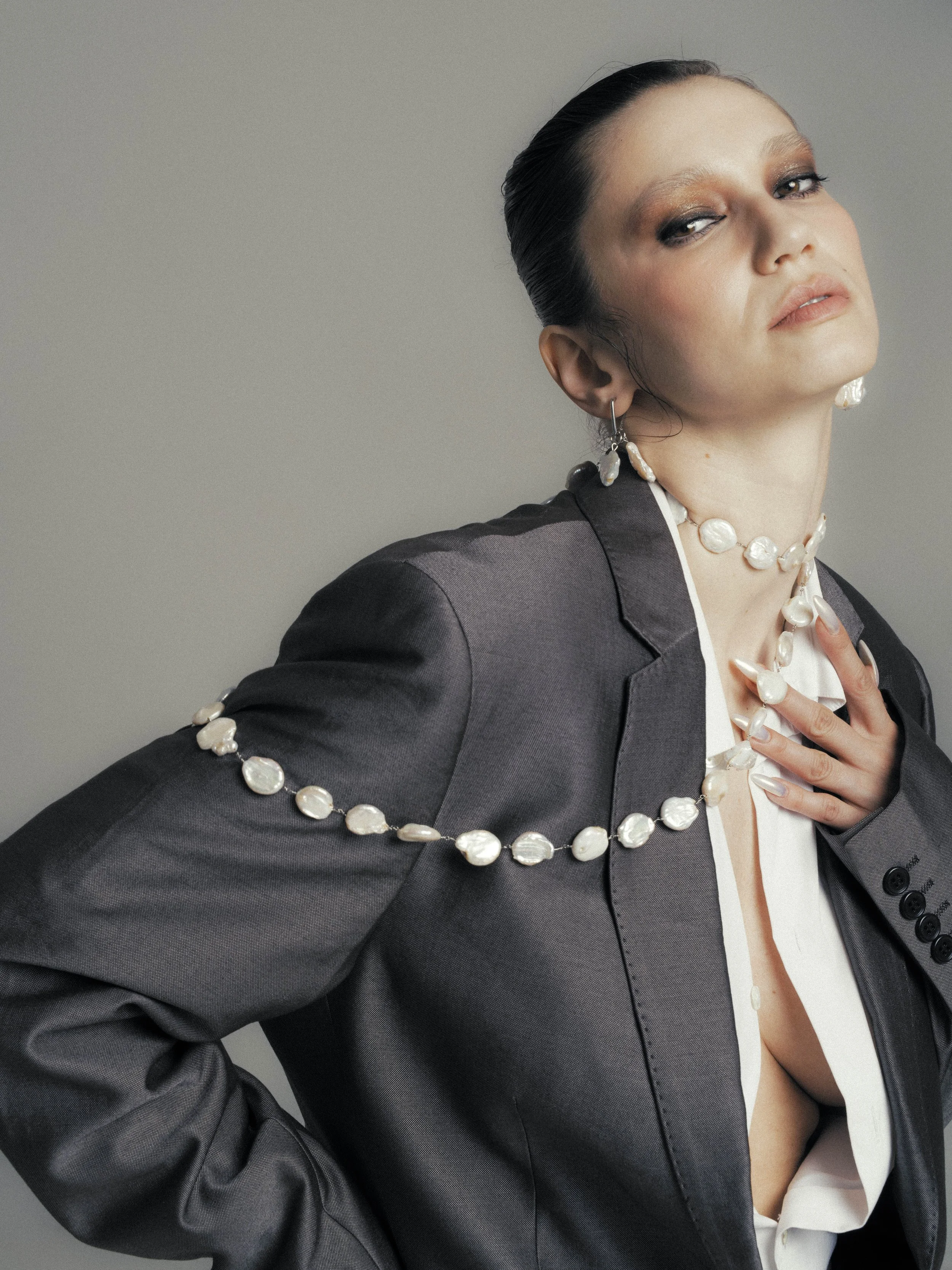 A woman with slicked-back dark hair wearing a dark suit jacket, white shirt, and pearl jewelry on her ear and neck, posing against a plain background.