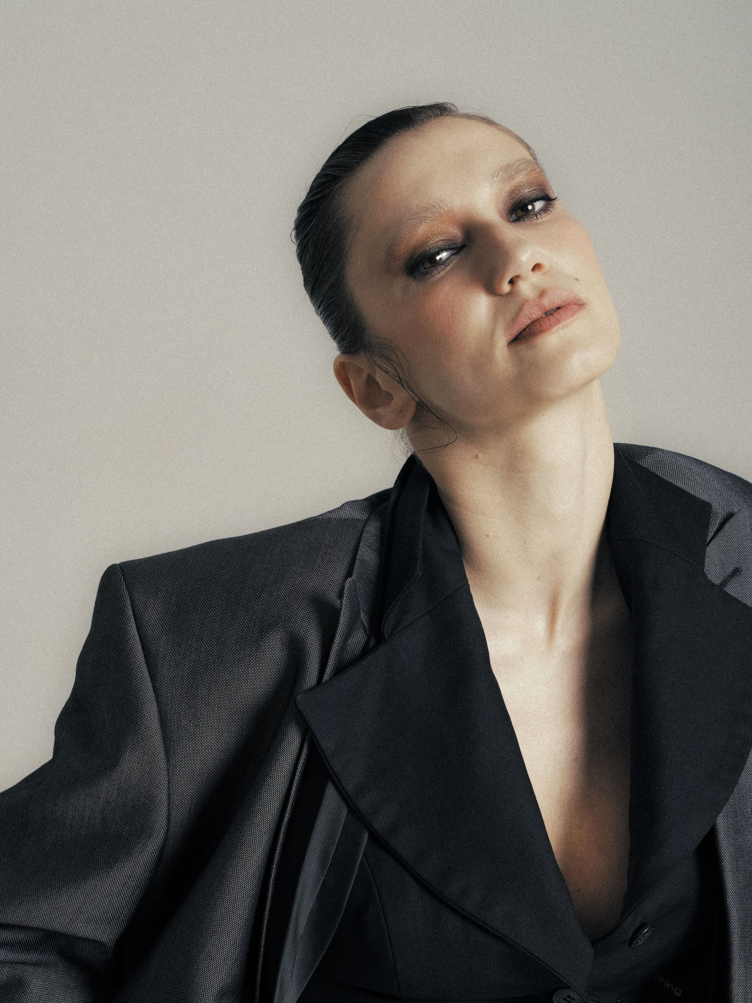 A woman with slicked back dark hair wearing a black blazer with sheer sleeves, posing against a plain white background.