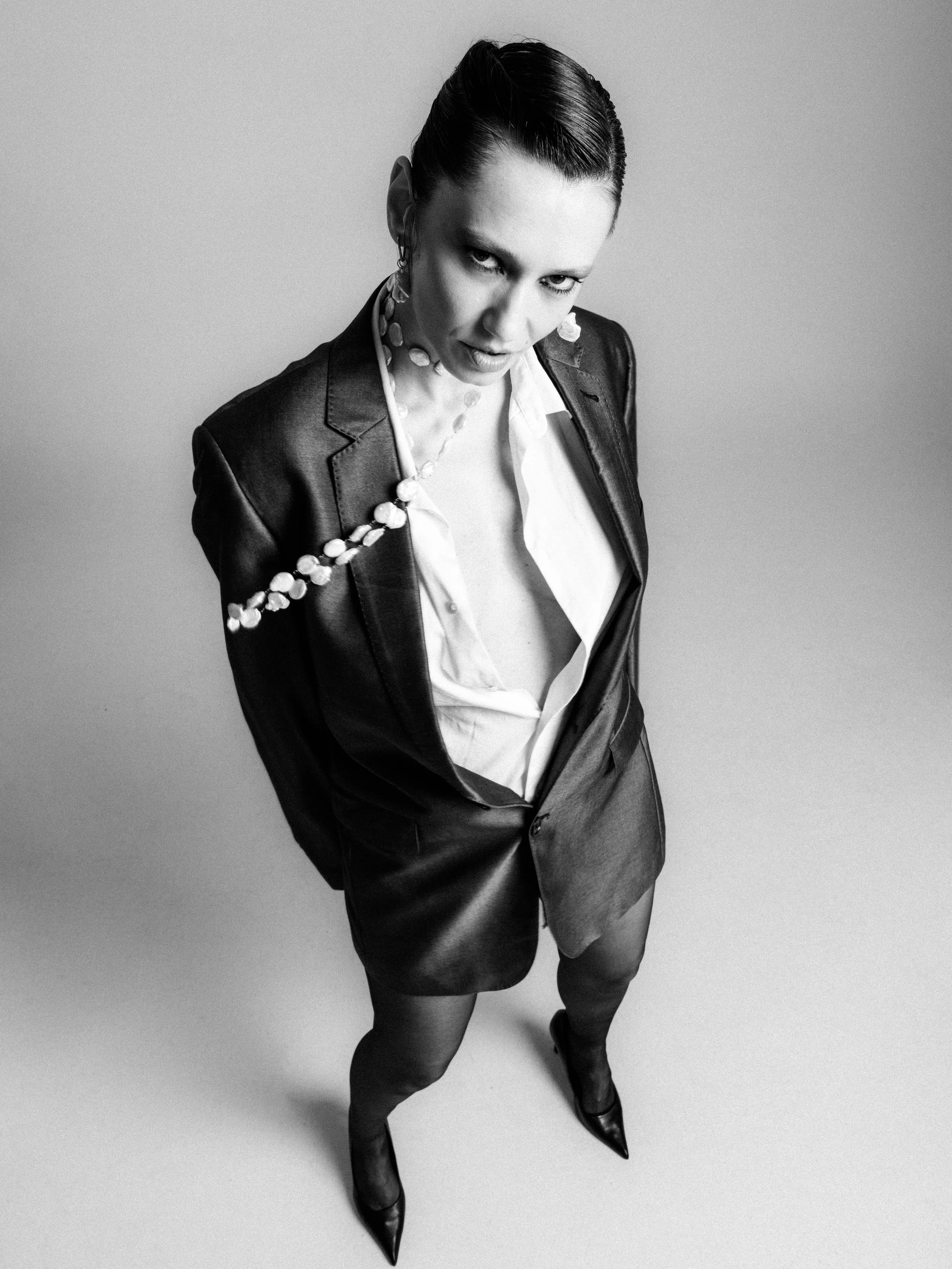 A woman in a leather blazer and skirt posing in black and white with short hair, pointing her toes in high heels, looking up at the camera.