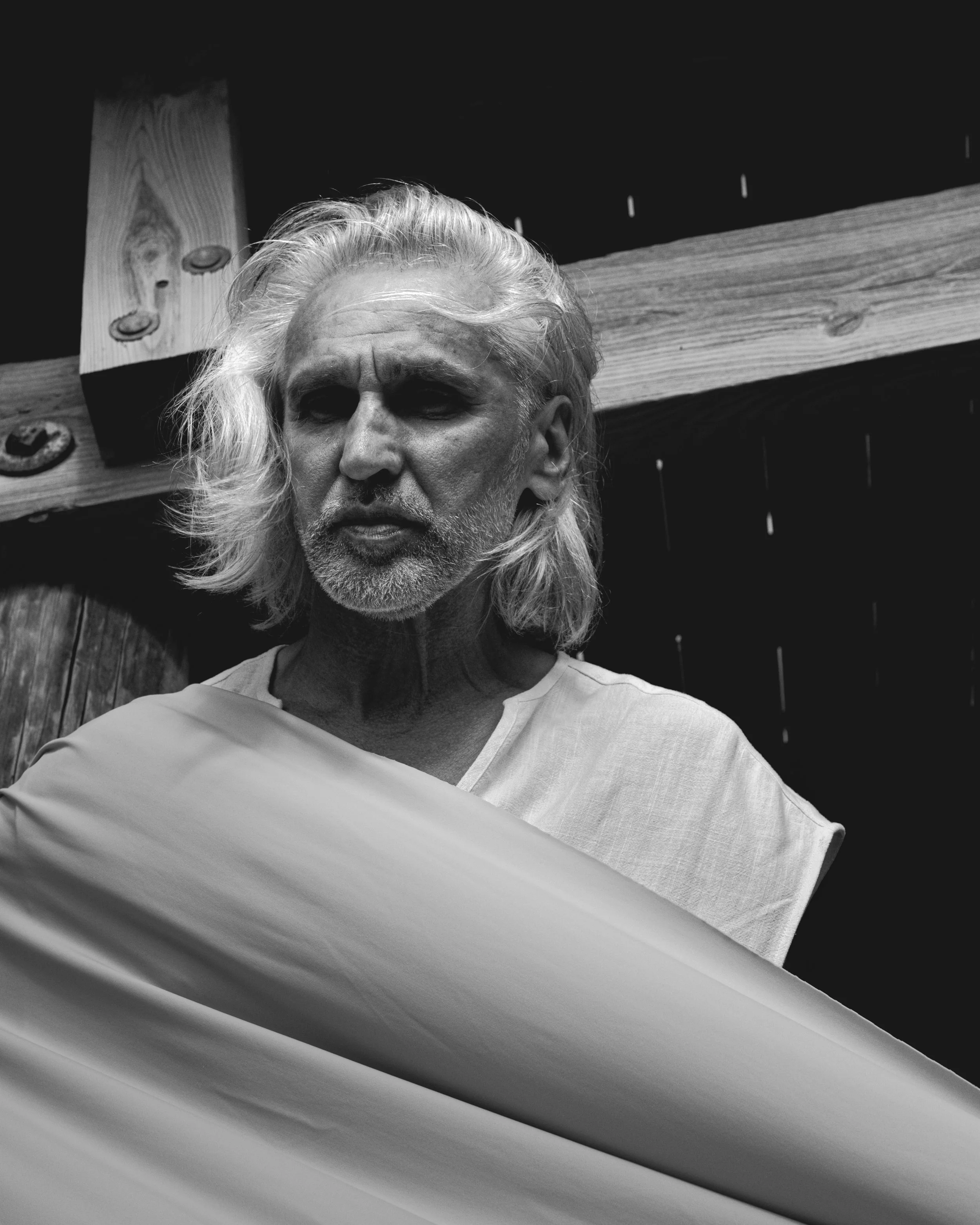 A man with long, wavy, gray hair and a beard wears a light-colored shirt and stands against wooden beams, some of which have nails. The photo is taken indoors or in a covered area in black and white.