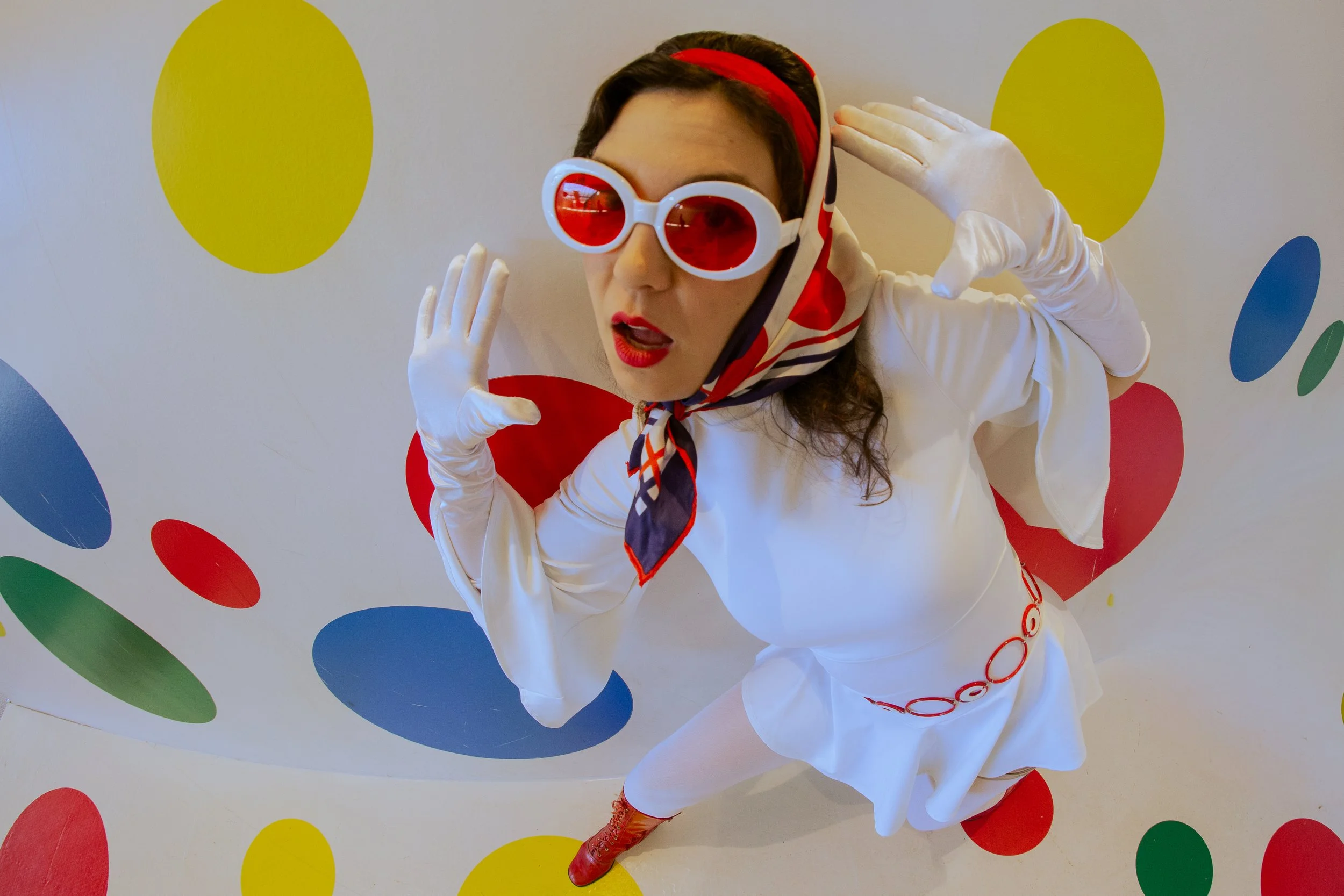 Person dressed in a white outfit with red accessories, wearing red sunglasses and a red and white headscarf, standing against a background with colorful polka dots.