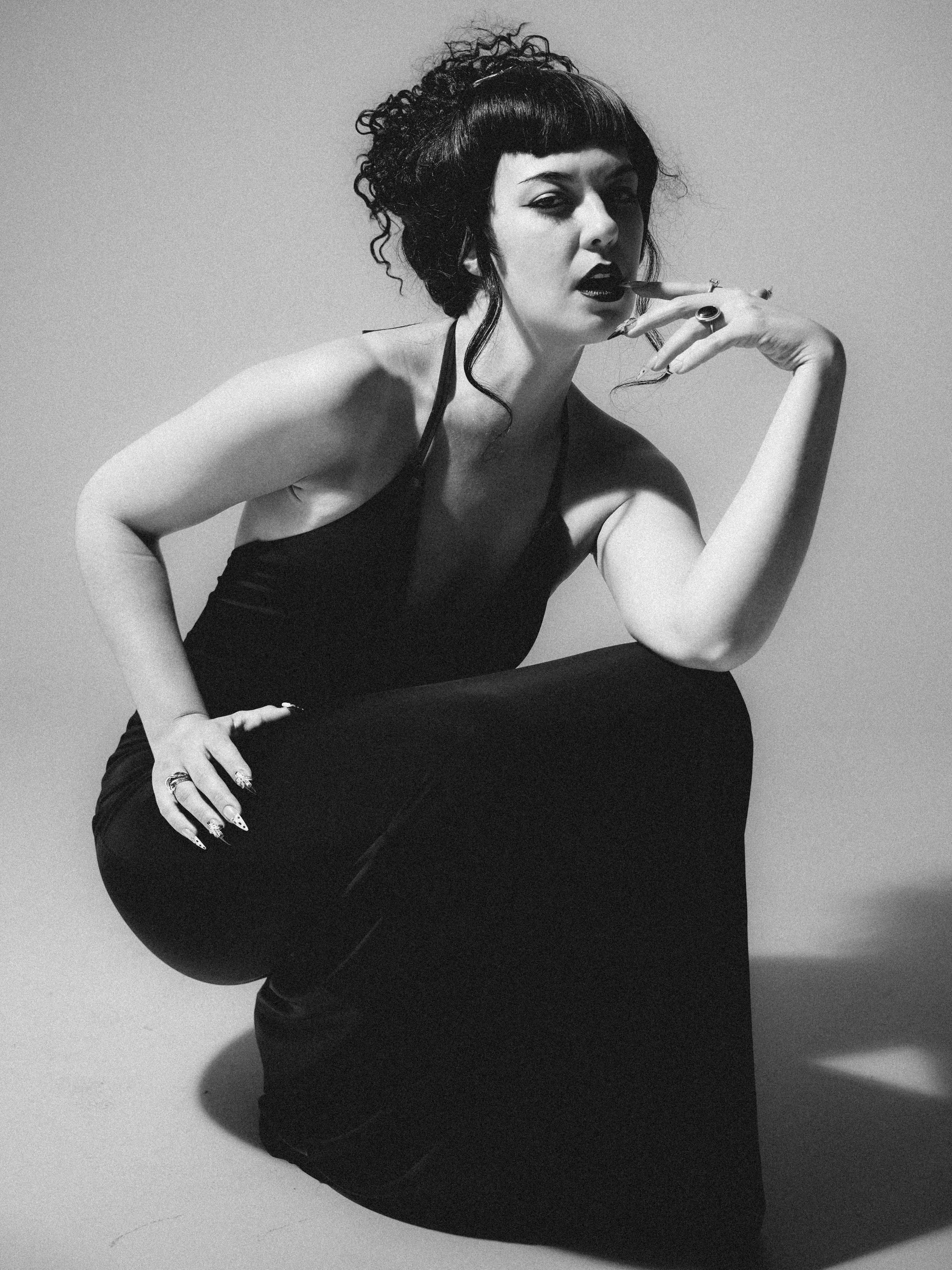 Black and white photo of a woman with dark lipstick and curly hair, sitting on the ground with one knee up. She is wearing a sleeveless top and has rings on her fingers, showing her nails and rings. She is touching her face with her hand, looking dir