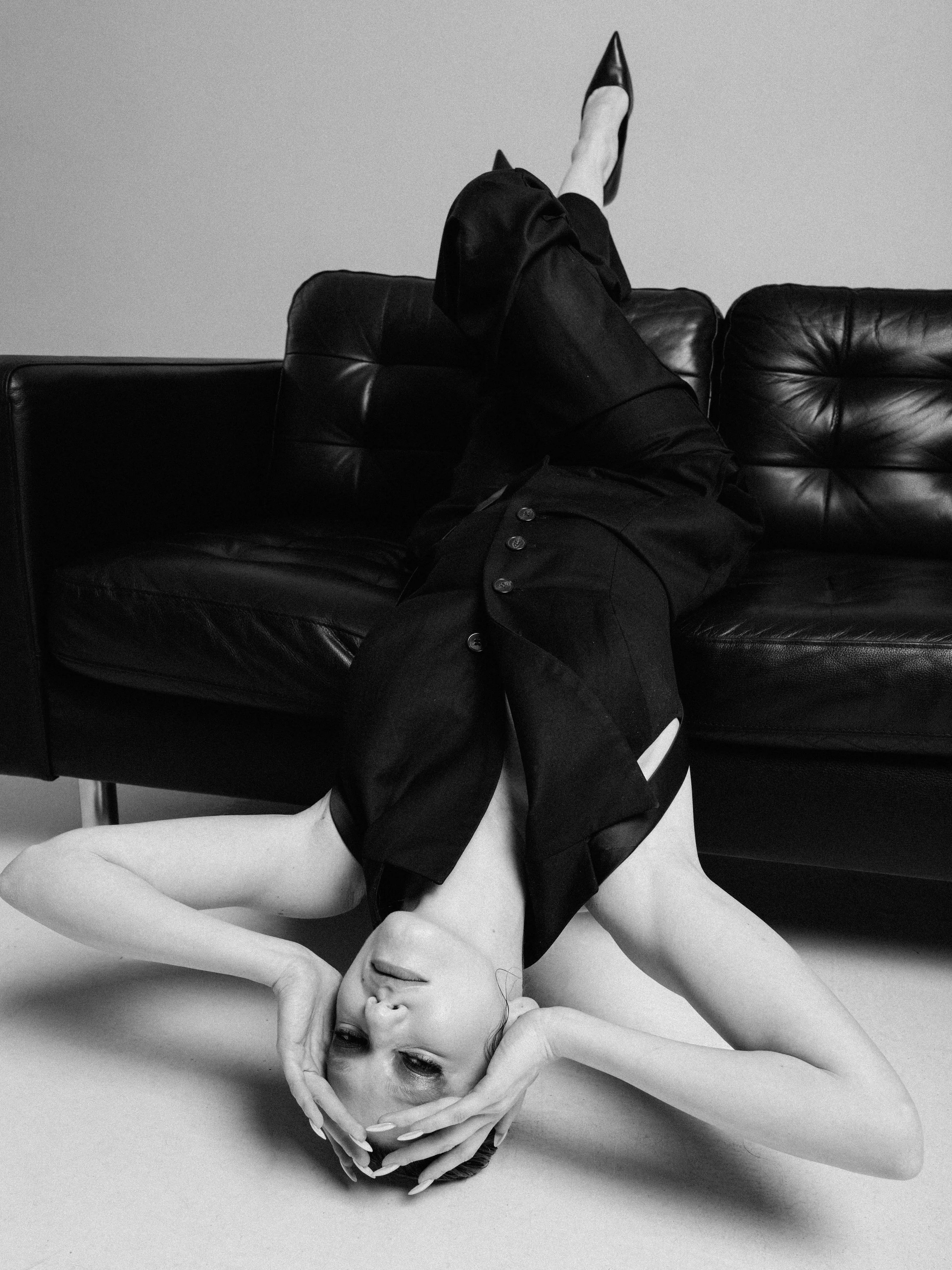 A black and white photograph of a woman lying on a surface, wearing a black sleeveless top and dark pants, with her head tilted back and her hand resting on her forehead. She has a relaxed expression and her eyes are closed.