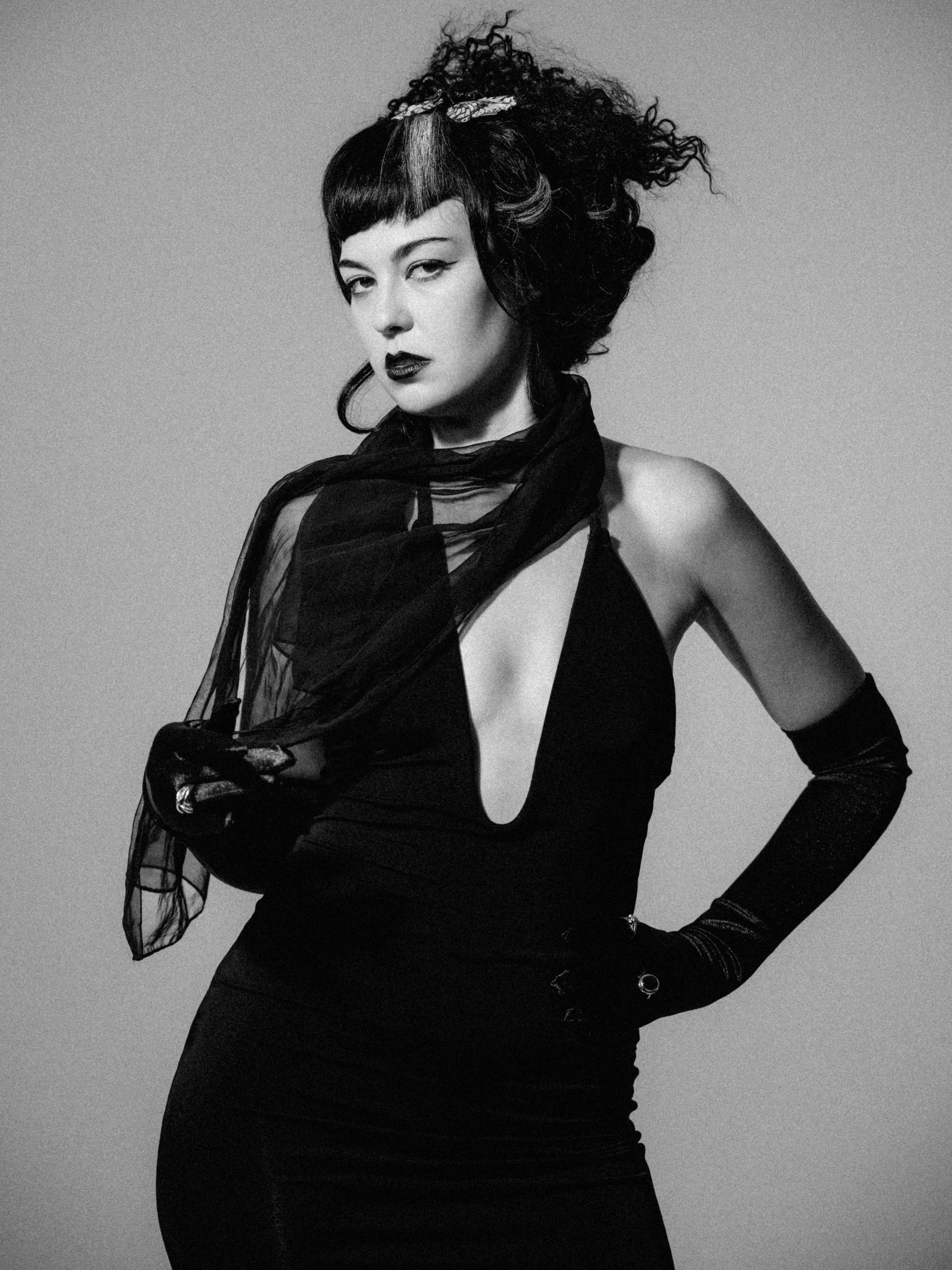 Black and white photo of a woman with dark lipstick, wearing a black dress with a cutout, and long black gloves. She has a voluminous hairstyle with curls and a bandana, striking a confident pose.