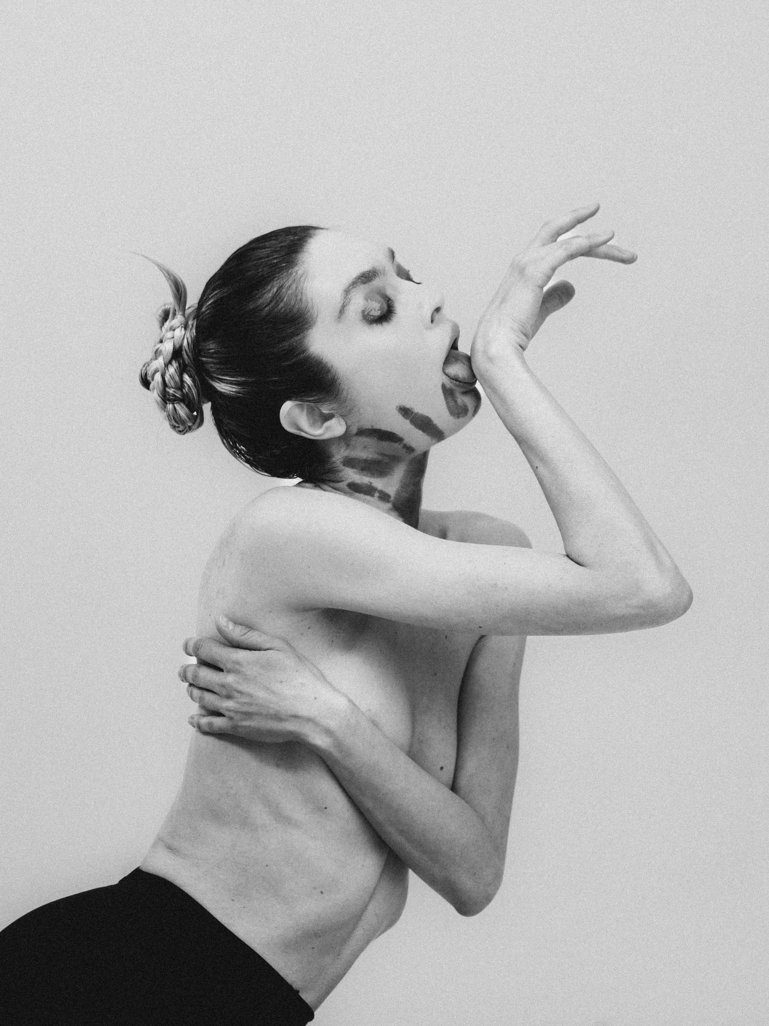 A woman with her hair in a braided bun sticking out her tongue and touching her finger to her lips, with distinct makeup and artistic markings on her neck and face, posing against a plain background.