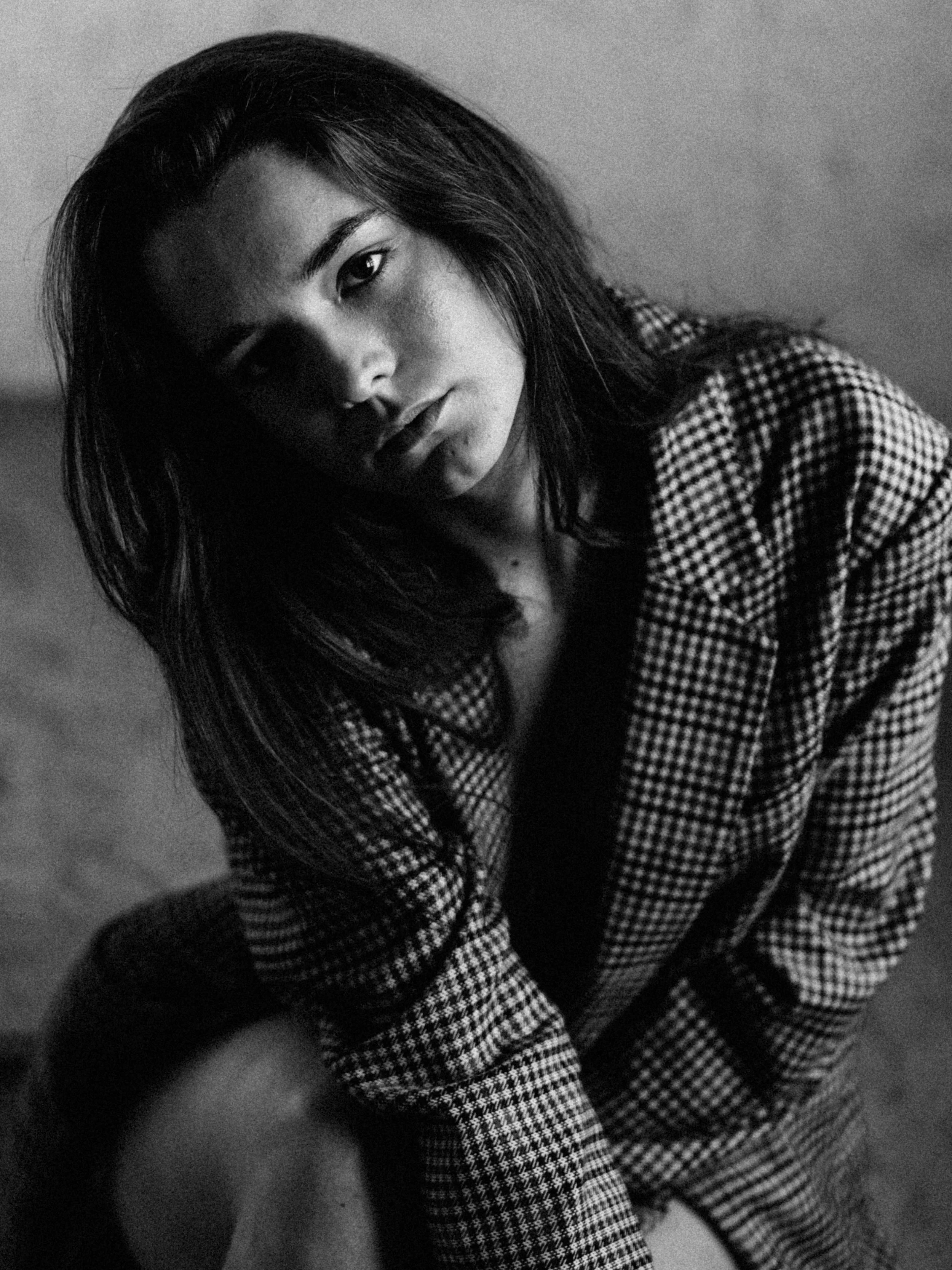 Black and white photo of a young woman with long dark hair, wearing a checkered top, looking directly at the camera with a serious expression.