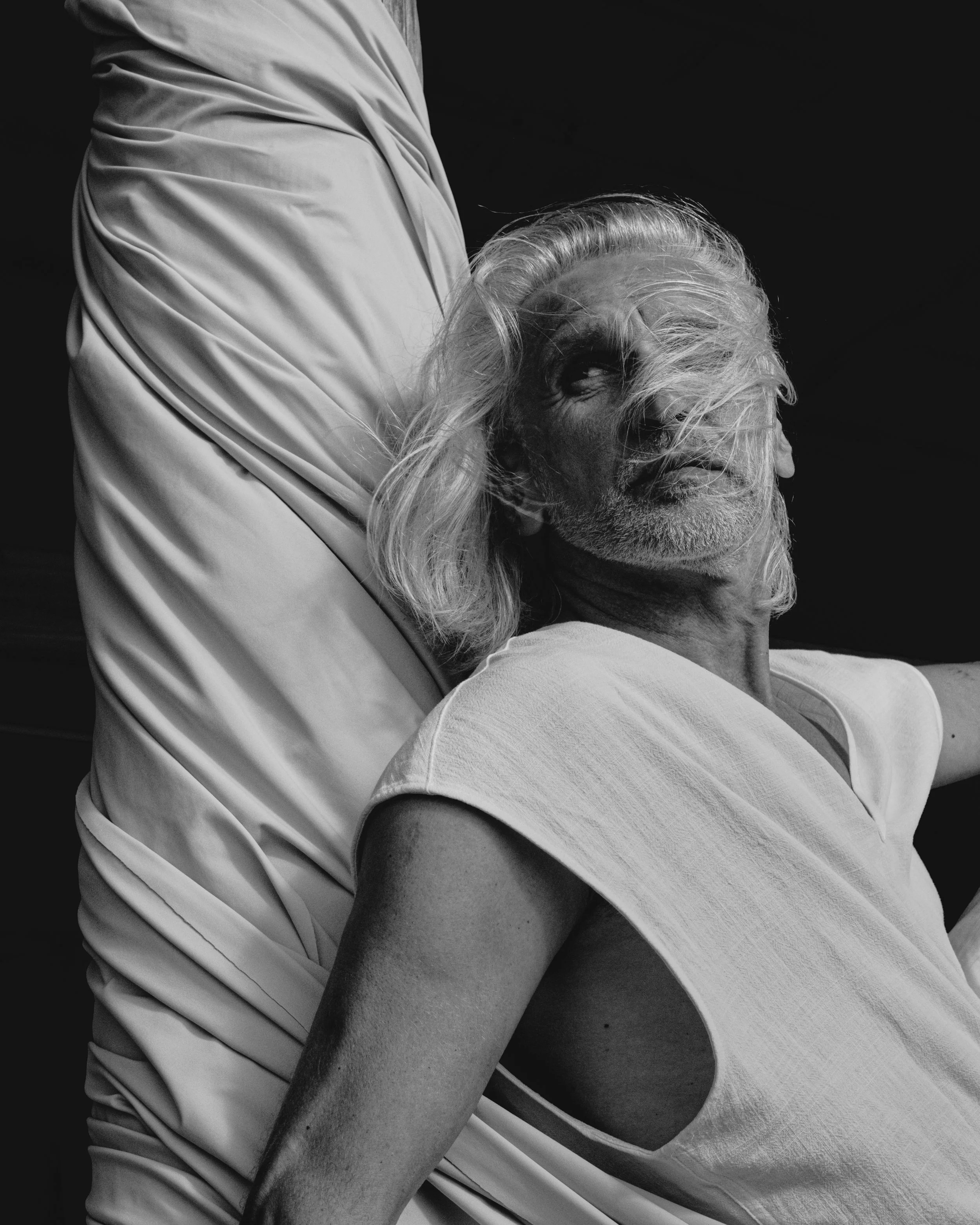 A black and white photo of a person with long white hair, sitting and resting their head on another person’s arm. The person with long hair is looking slightly upward with a relaxed expression, wearing a light-colored shirt. The other person’s arm is