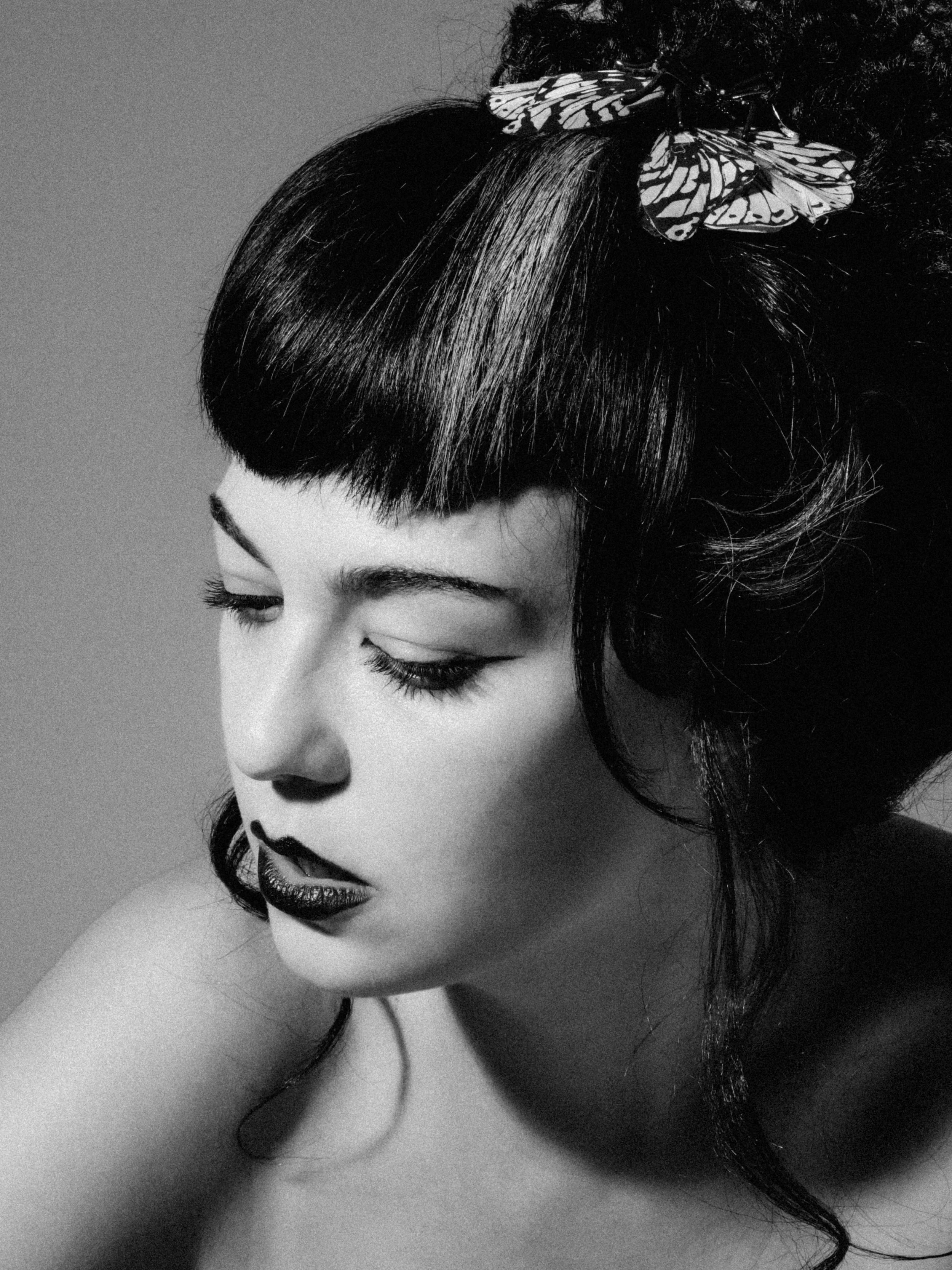 Black and white portrait of a woman with styled hair, including a butterfly hair accessory, with makeup and closed eyes.