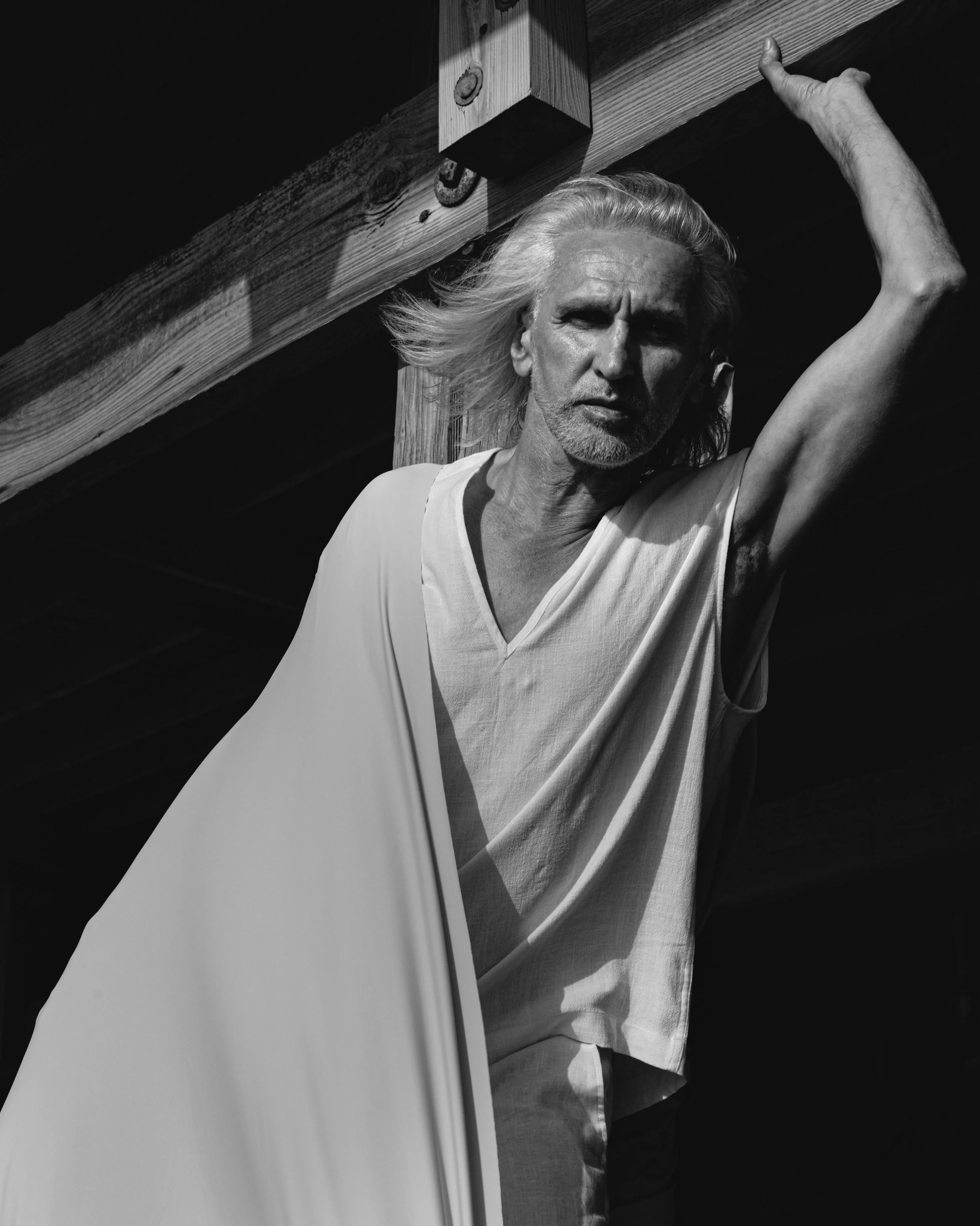 Black and white photo of a man with long, flowing hair and a beard, standing under a wooden structure with his arm raised, looking directly at the camera.