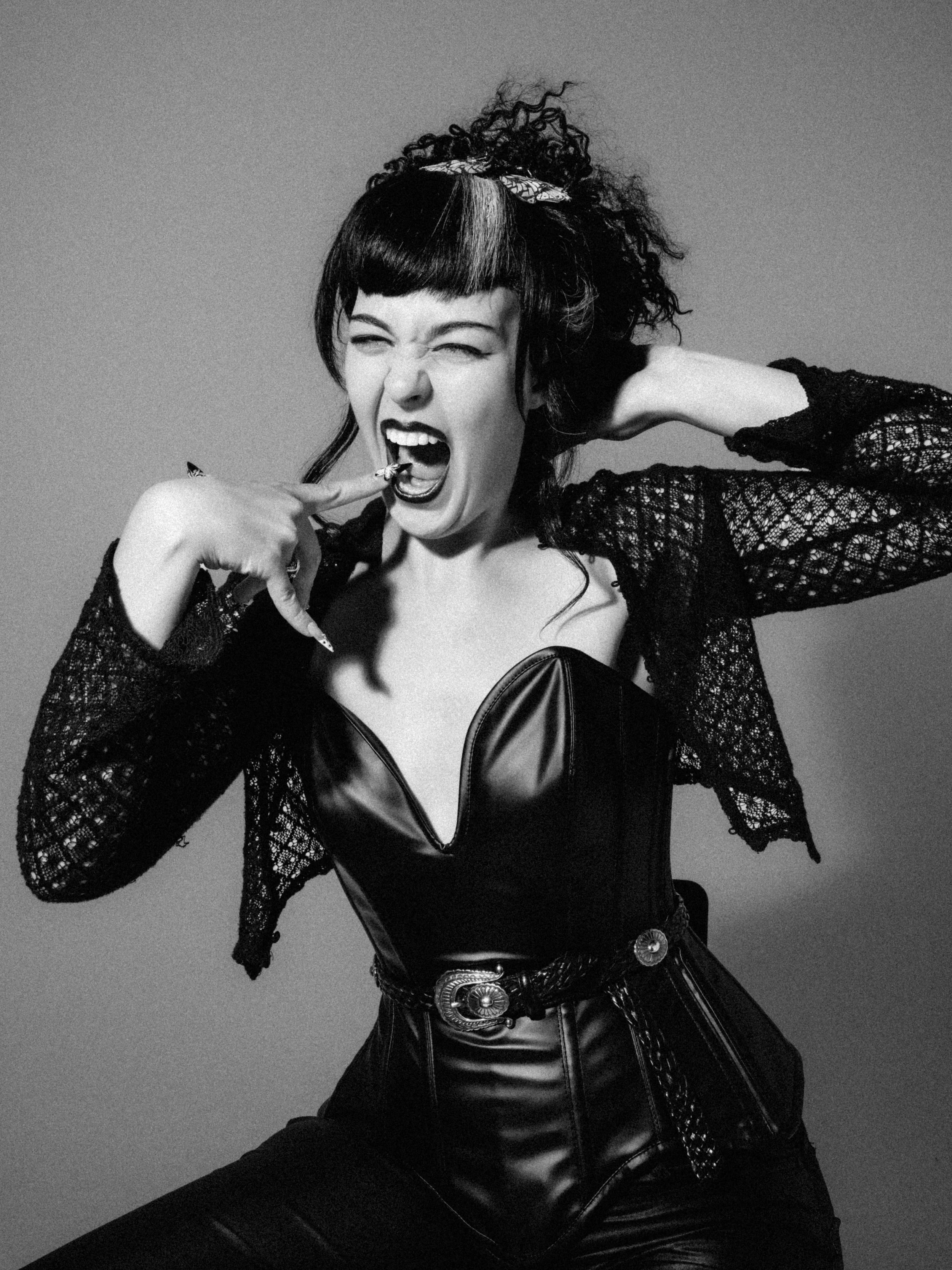 Black and white photo of a woman with curly and straight hair, wearing a leather corset, lace jacket, and belt with decorative buckles, making an expressive facial gesture with one eye squinted and her mouth open, touching her face with her finger.