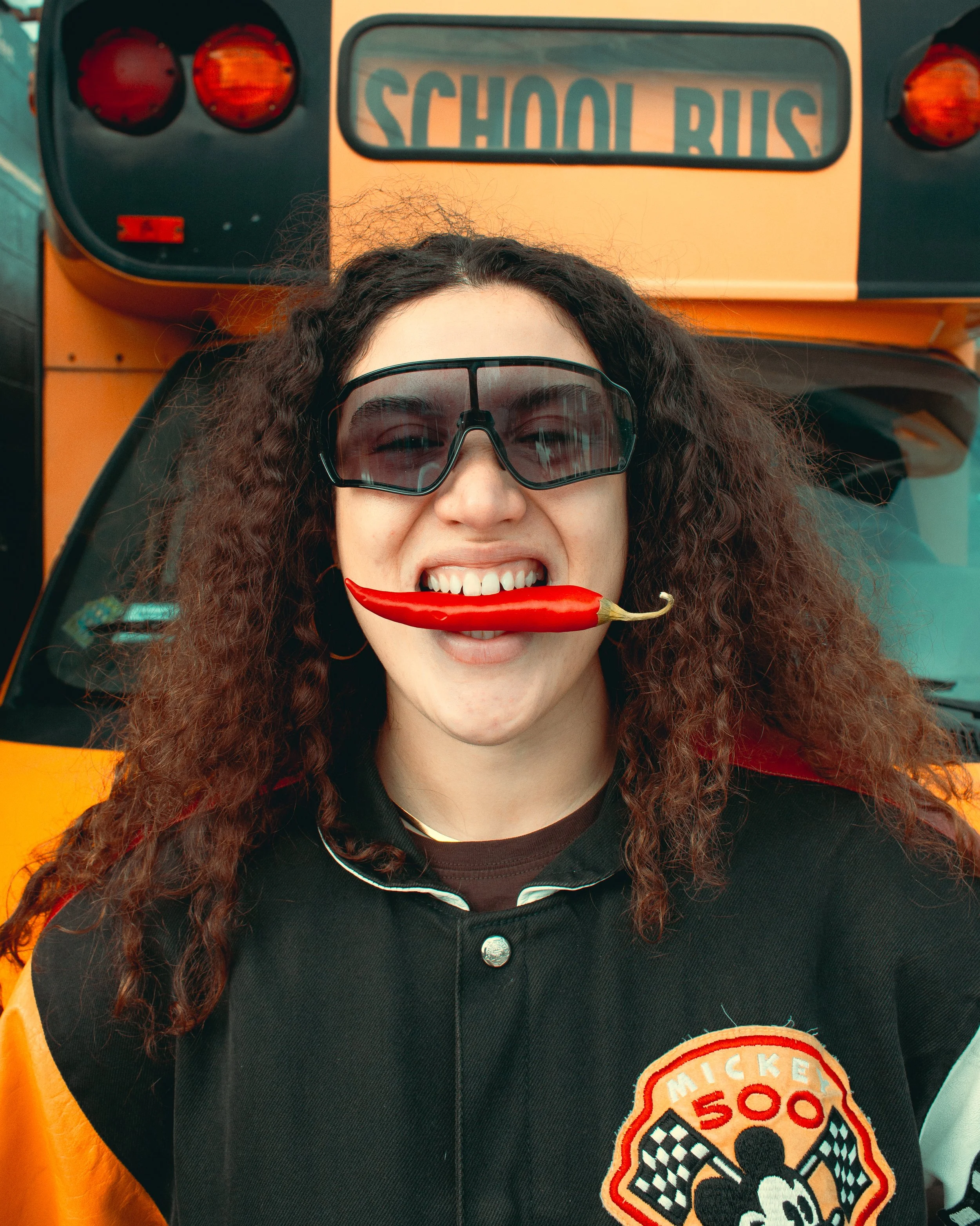 A woman with curly hair and sunglasses smiling while holding a red chili pepper in her mouth, standing in front of a yellow school bus that has a 'SCHOOL BUS' sign on the back. She is wearing a black shirt with a Mickey Mouse embroidered patch.