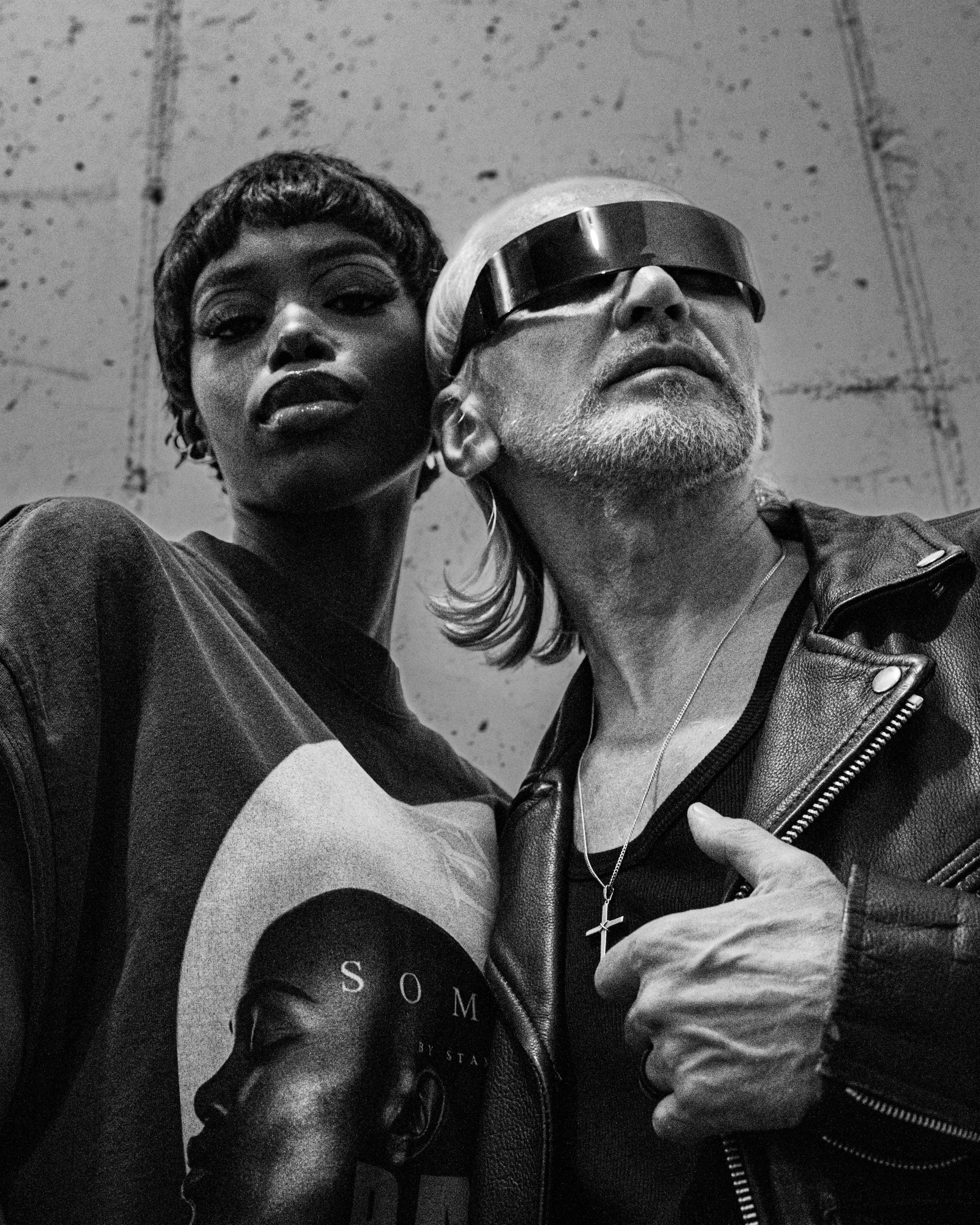 A black and white photo of a woman with short hair and dark makeup next to an older man with gray hair and a beard, wearing large futuristic glasses. They are close together, with the woman's arm partially visible taking a selfie.