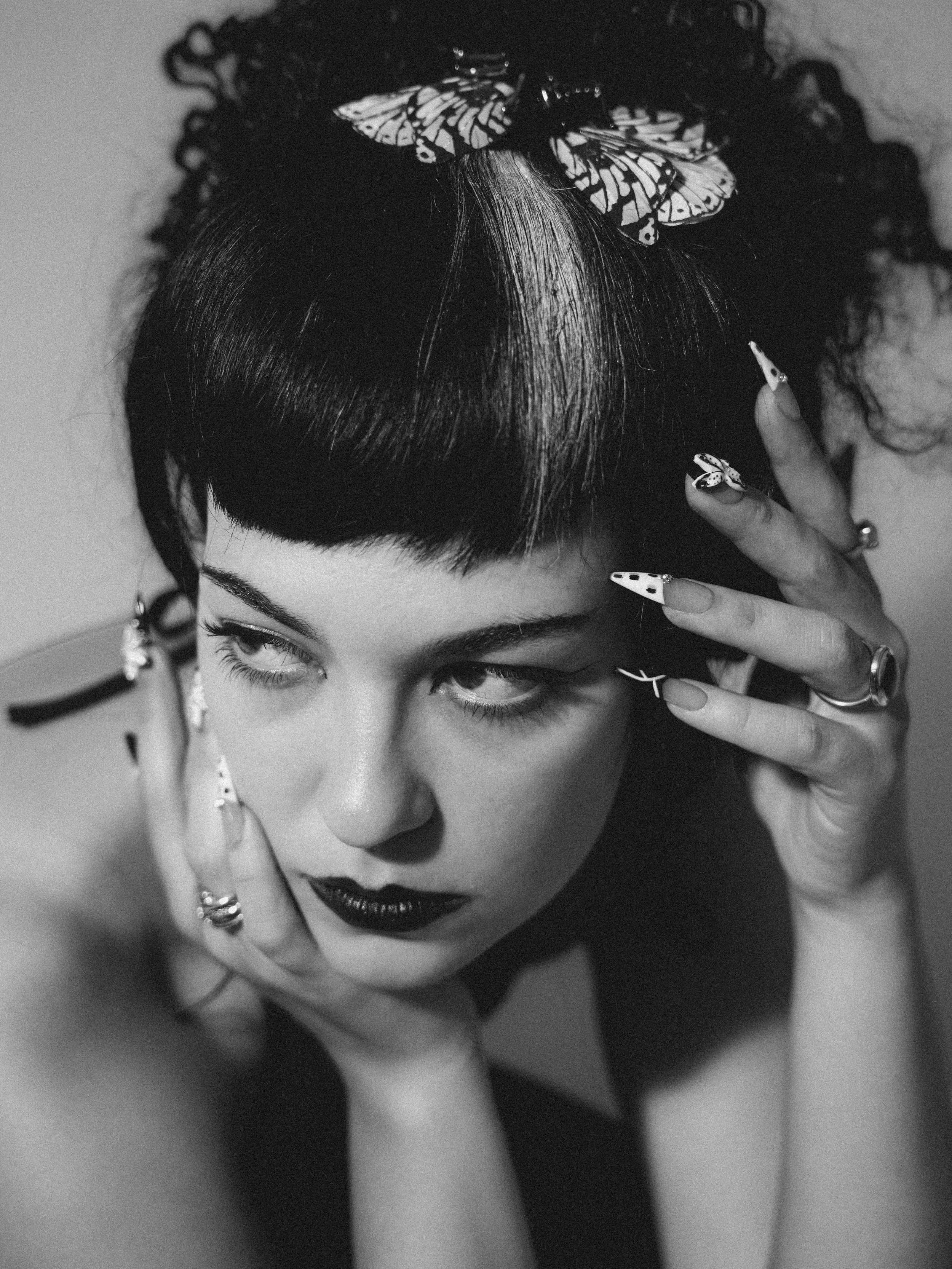 A black and white photograph of a woman with dark lipstick and eye makeup, holding her face with her hand. She has nail art and rings, and butterflies on her head and hand.