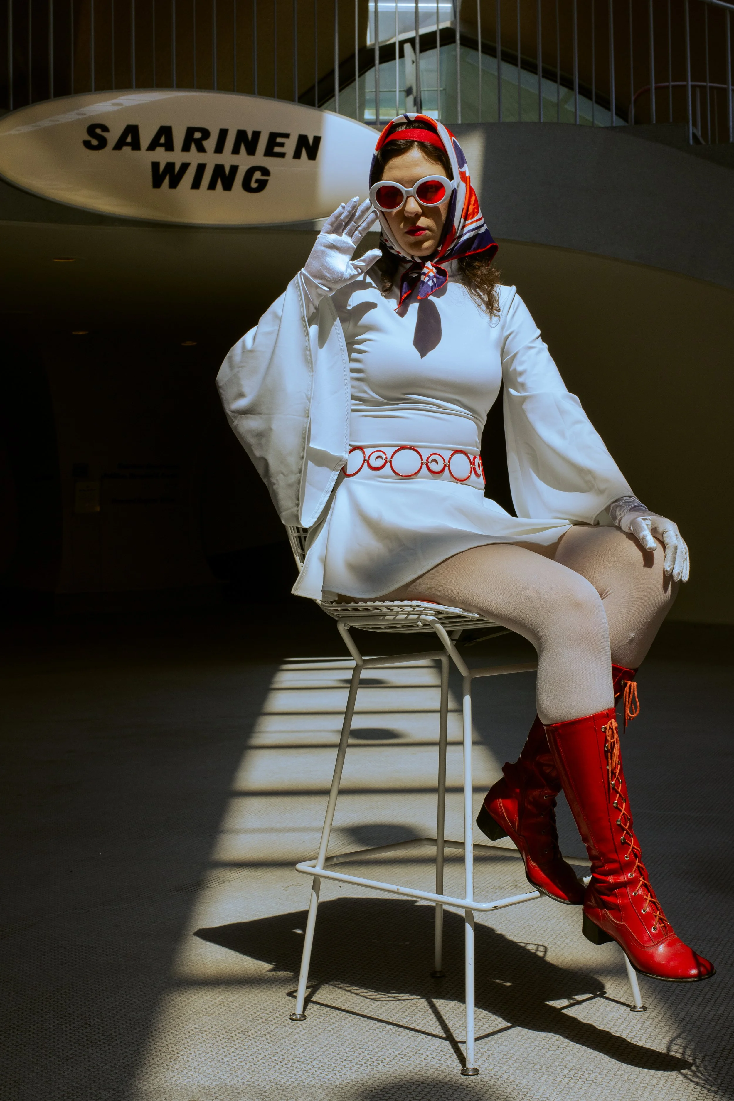 Woman in vintage flight attendant outfit, red leather lace-up boots, white dress, large red and white sunglasses, and a headscarf with red, white, and blue colors, sitting on a white chair in front of an airport sign reading "Saarinen Wing".