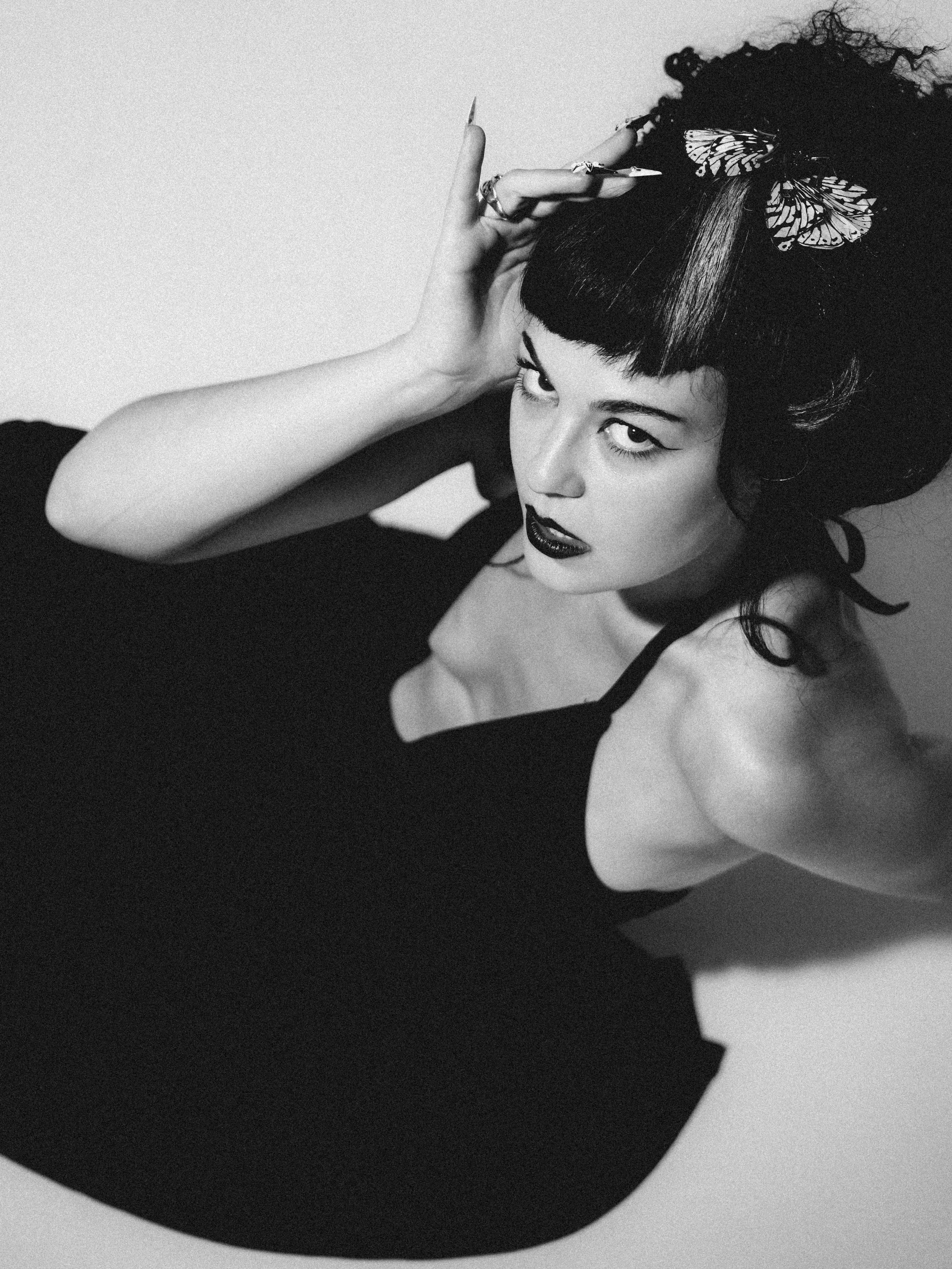 A black-and-white photograph of a woman with short hair, dark lipstick, and eyeliner in a dramatic pose with butterfly hair accessories.