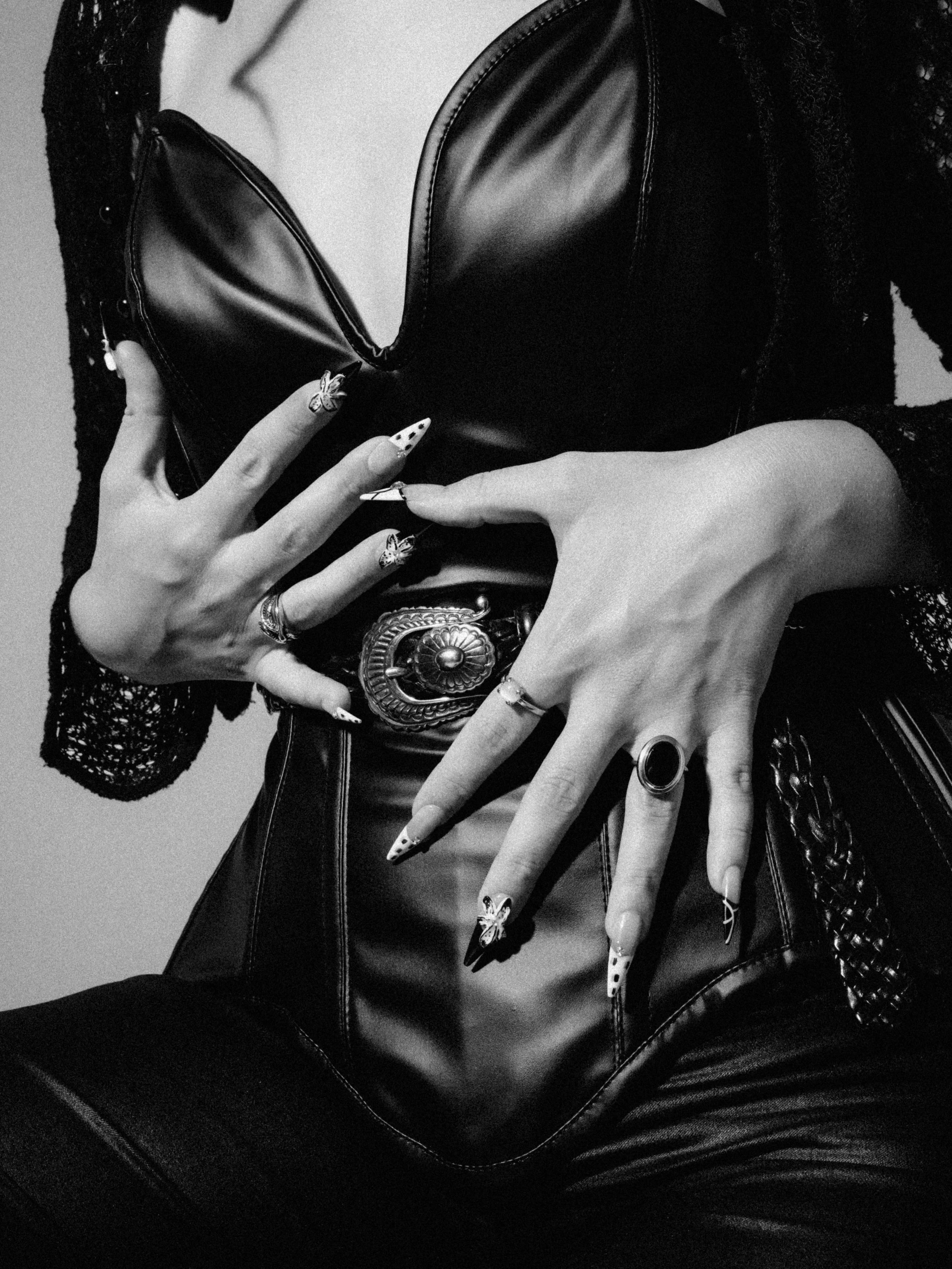 Person wearing a leather jacket and lace sleeves, holding a clutch purse with tattoos on fingers, wearing rings and bracelets, with butterfly-shaped nails.