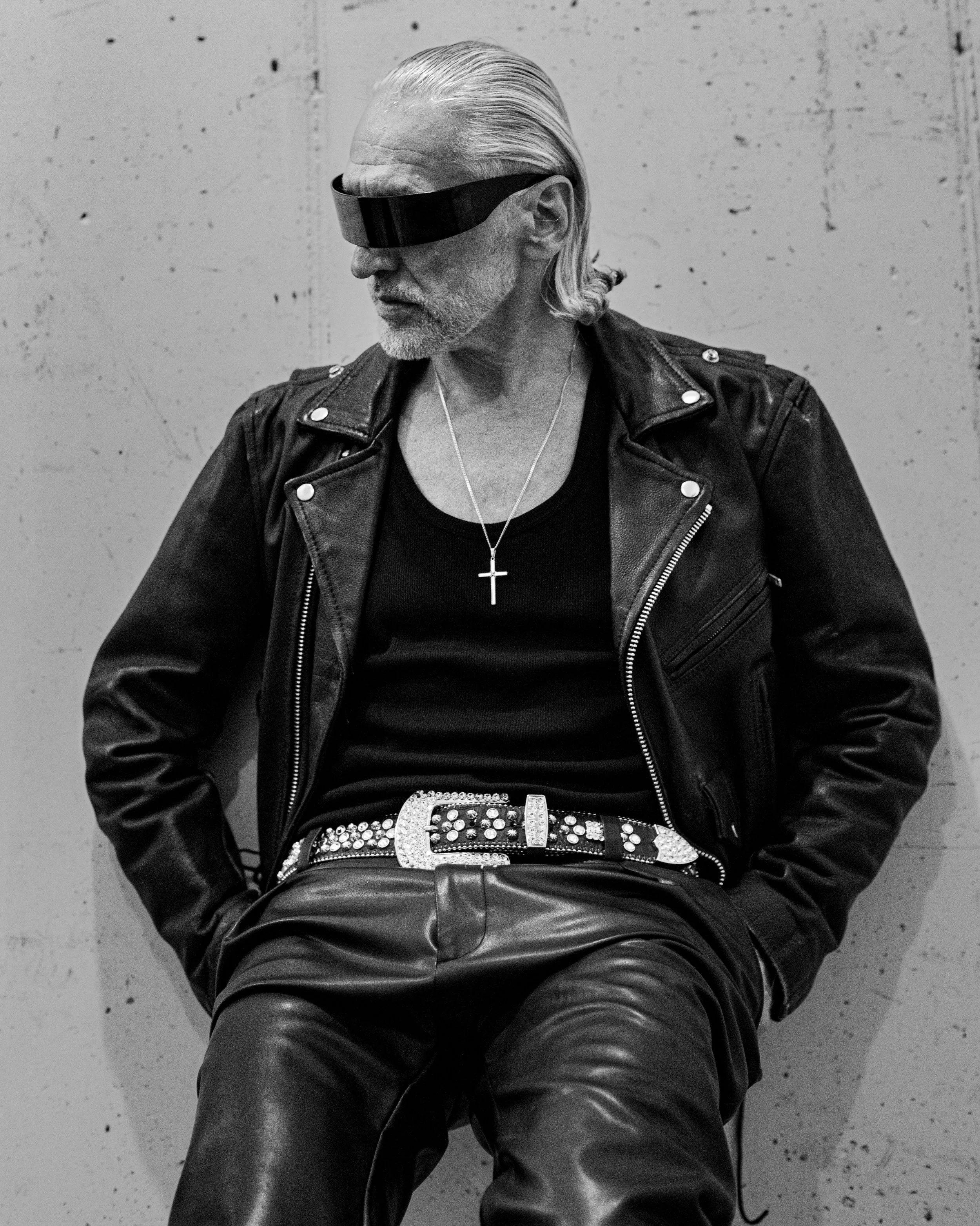 Black and white photograph of an older man with slicked-back hair and a beard wearing futuristic goggles, a leather jacket, a necklace with a cross pendant, and leather pants, sitting against a plain wall.