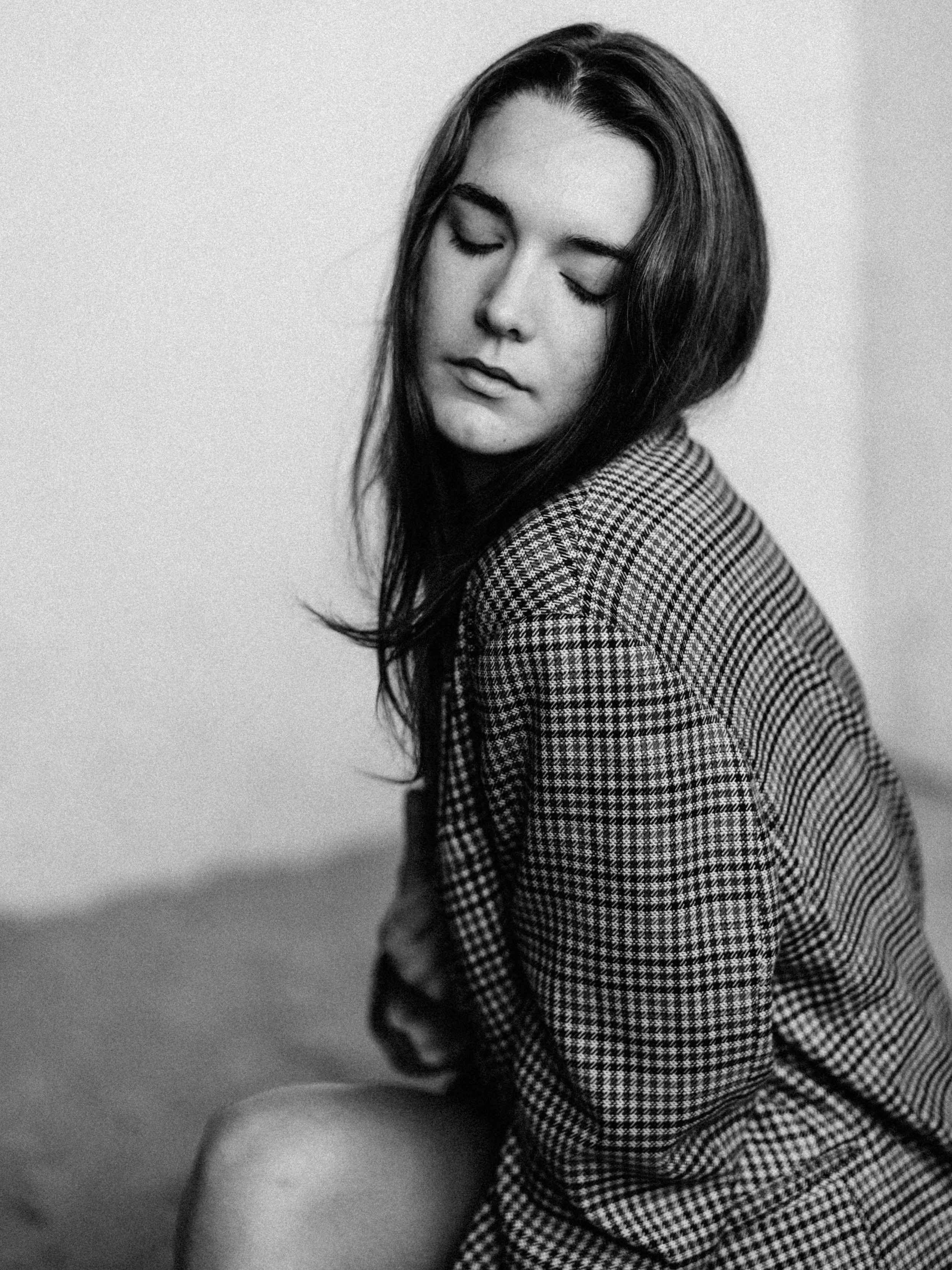 Black and white photo of a young woman with long hair, closed eyes, and a checkered shirt, looking down, sitting against a plain wall.
