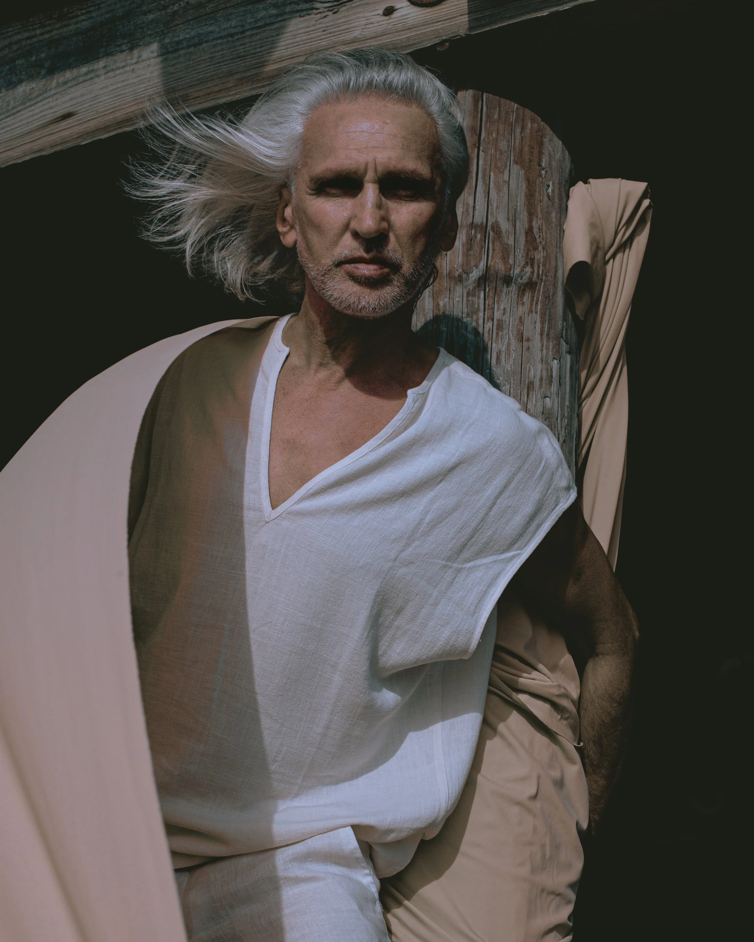 Head and shoulders of an older man with gray hair, wearing a white shirt, leaning against a wooden post outdoors, with another person's arm partially visible in the background.
