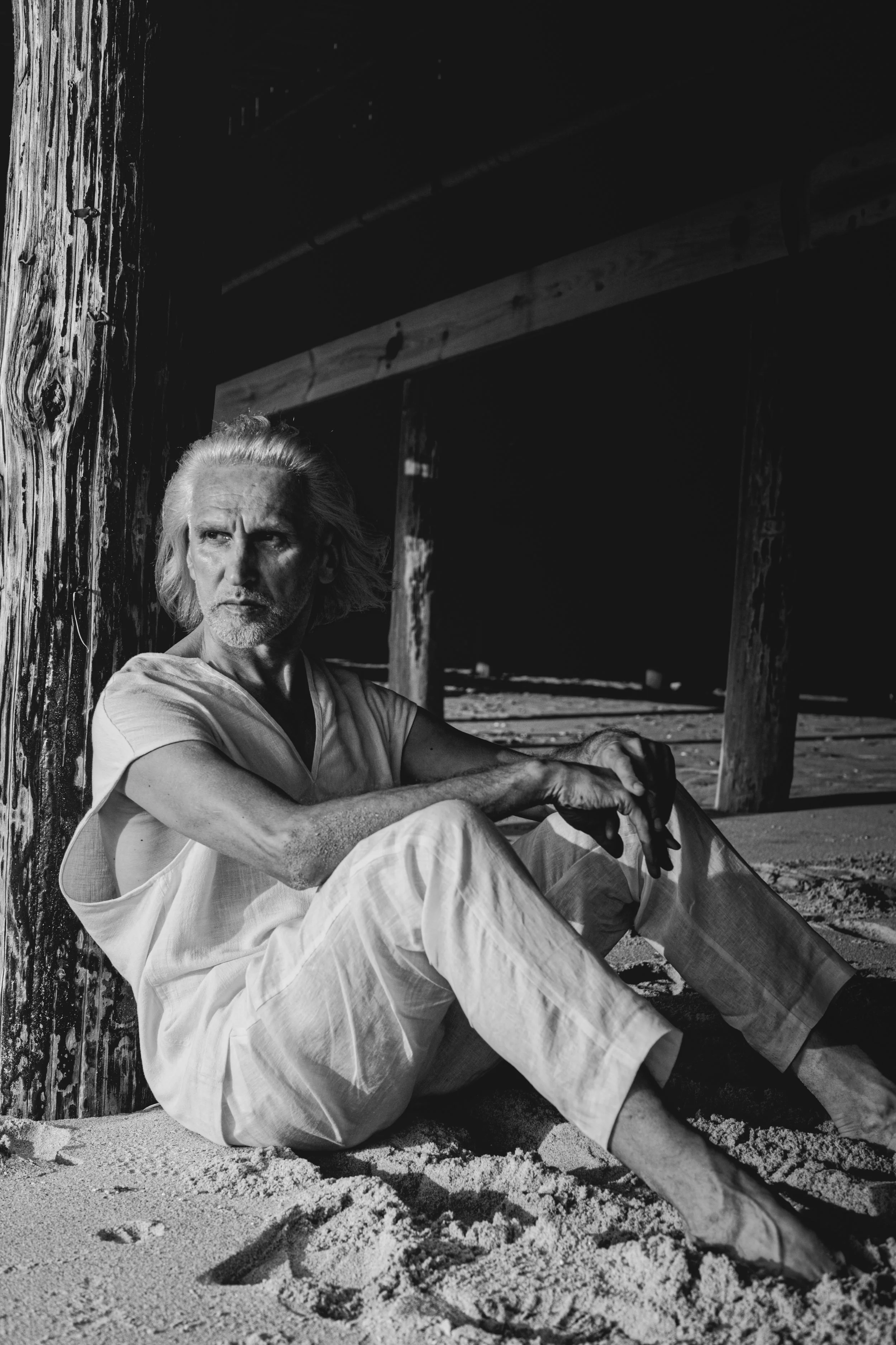 An older man with long, gray hair and a beard, wearing light-colored clothing, sitting on a sandy surface under a wooden structure, looking off to the side.