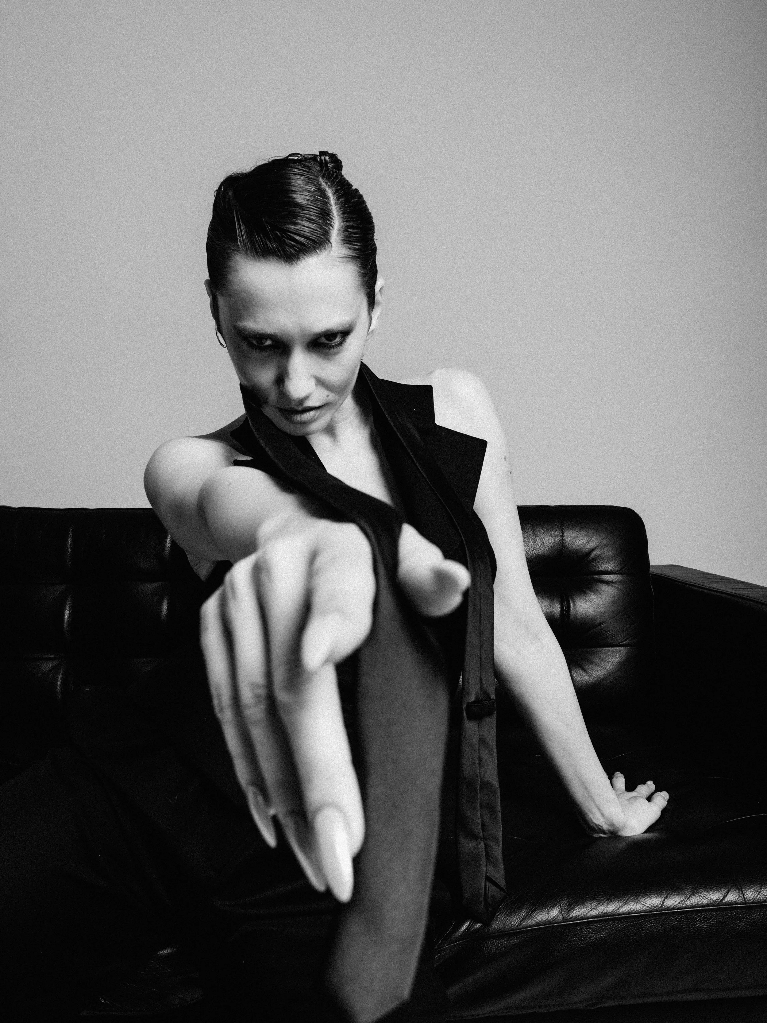 Black and white photo of a woman with slicked back hair, intense gaze, and pointing finger, seated on a black leather couch against a plain wall.