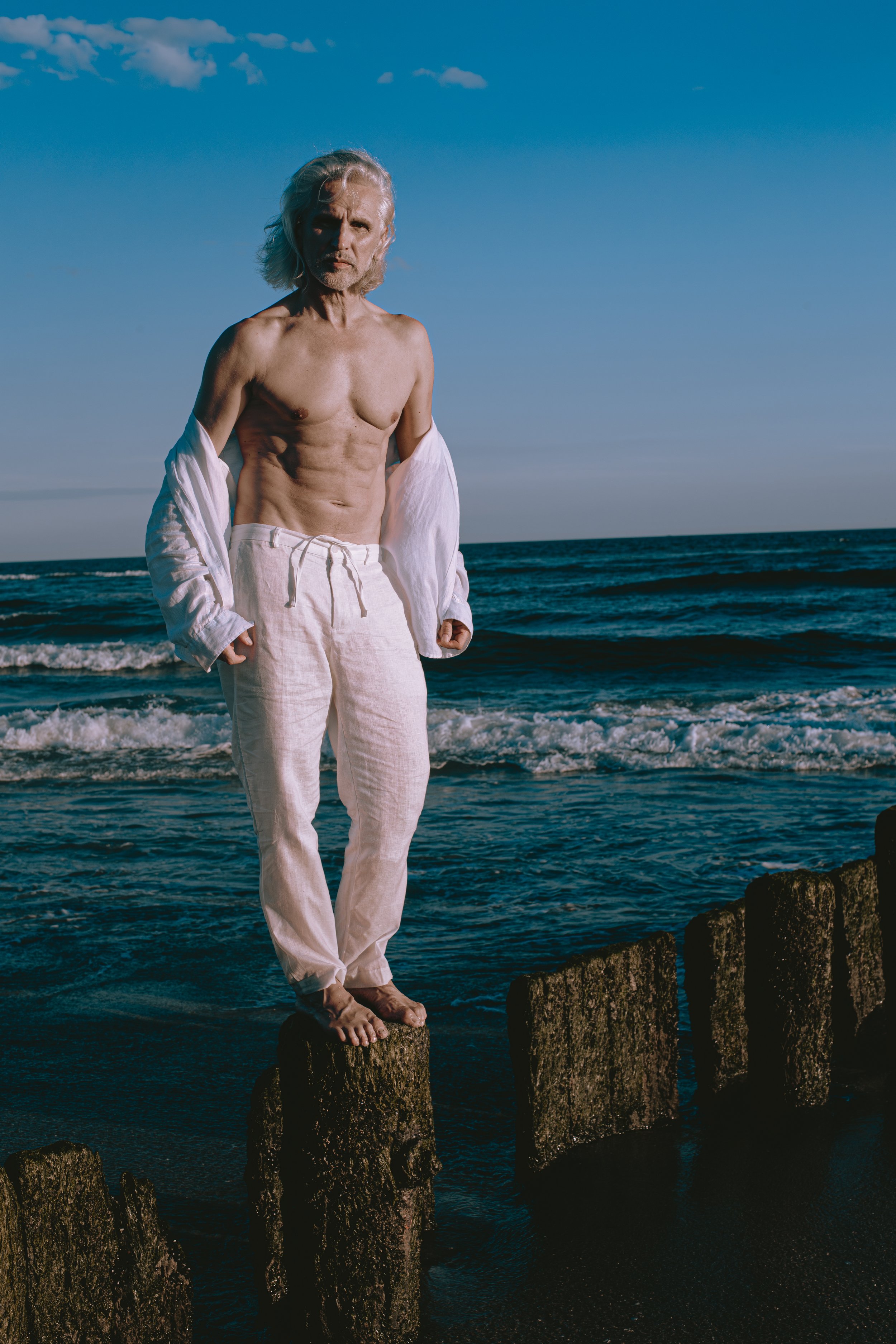 An elderly man with long gray hair standing on wooden posts at the beach during sunset, shirtless with a white open shirt and pants.