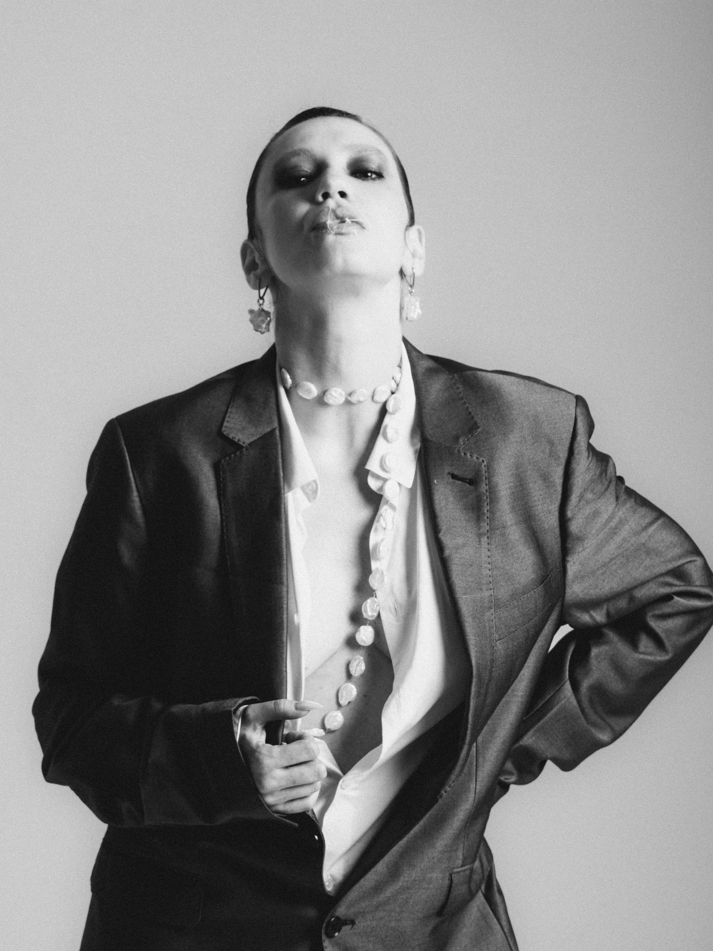 Black and white photograph of a woman wearing a leather blazer, white shirt with pearl buttons, and statement earrings, with her hair slicked back, looking confidently at the camera.