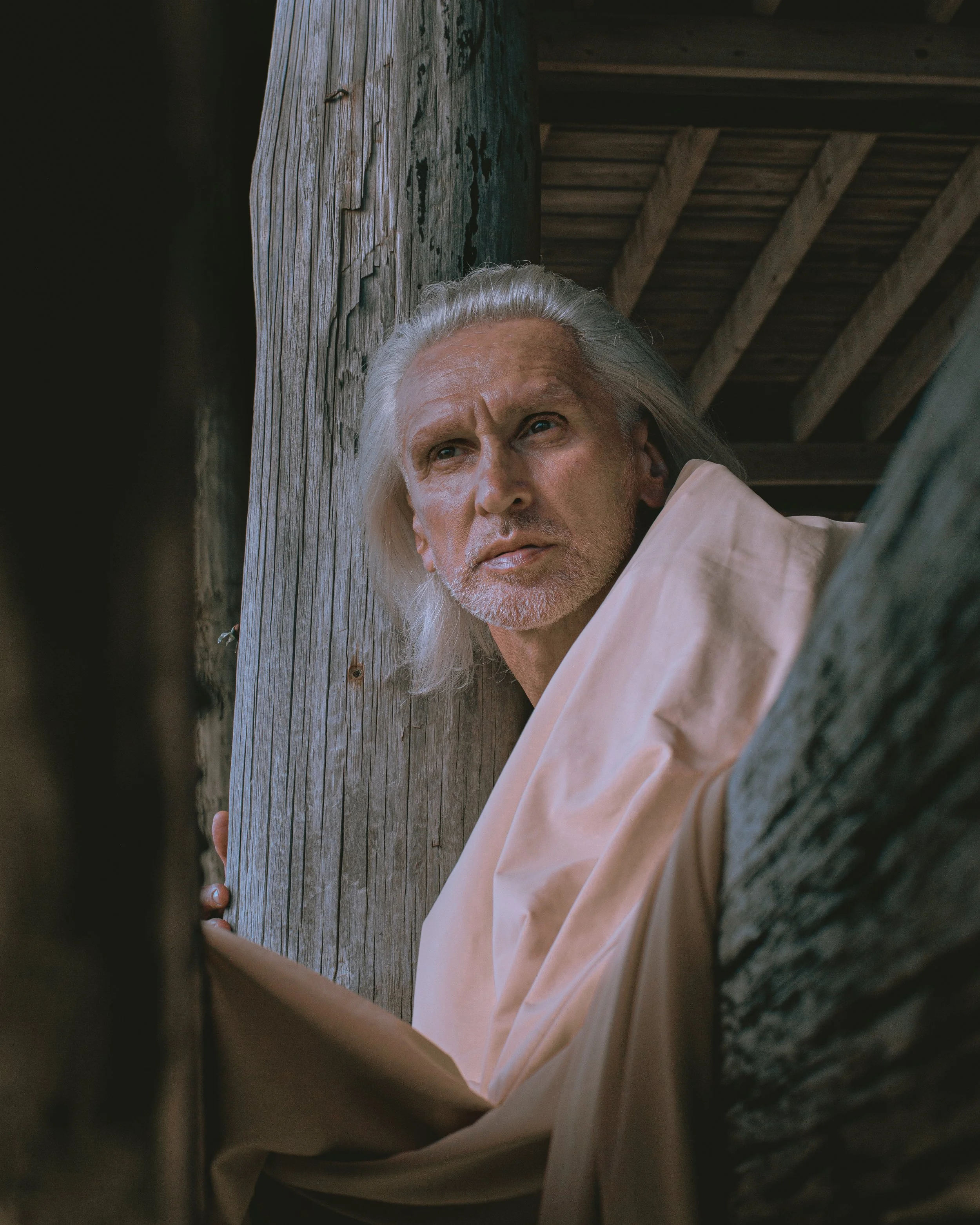 A man with long silver hair and a beard lies on the ground under a wooden structure, looking upwards.