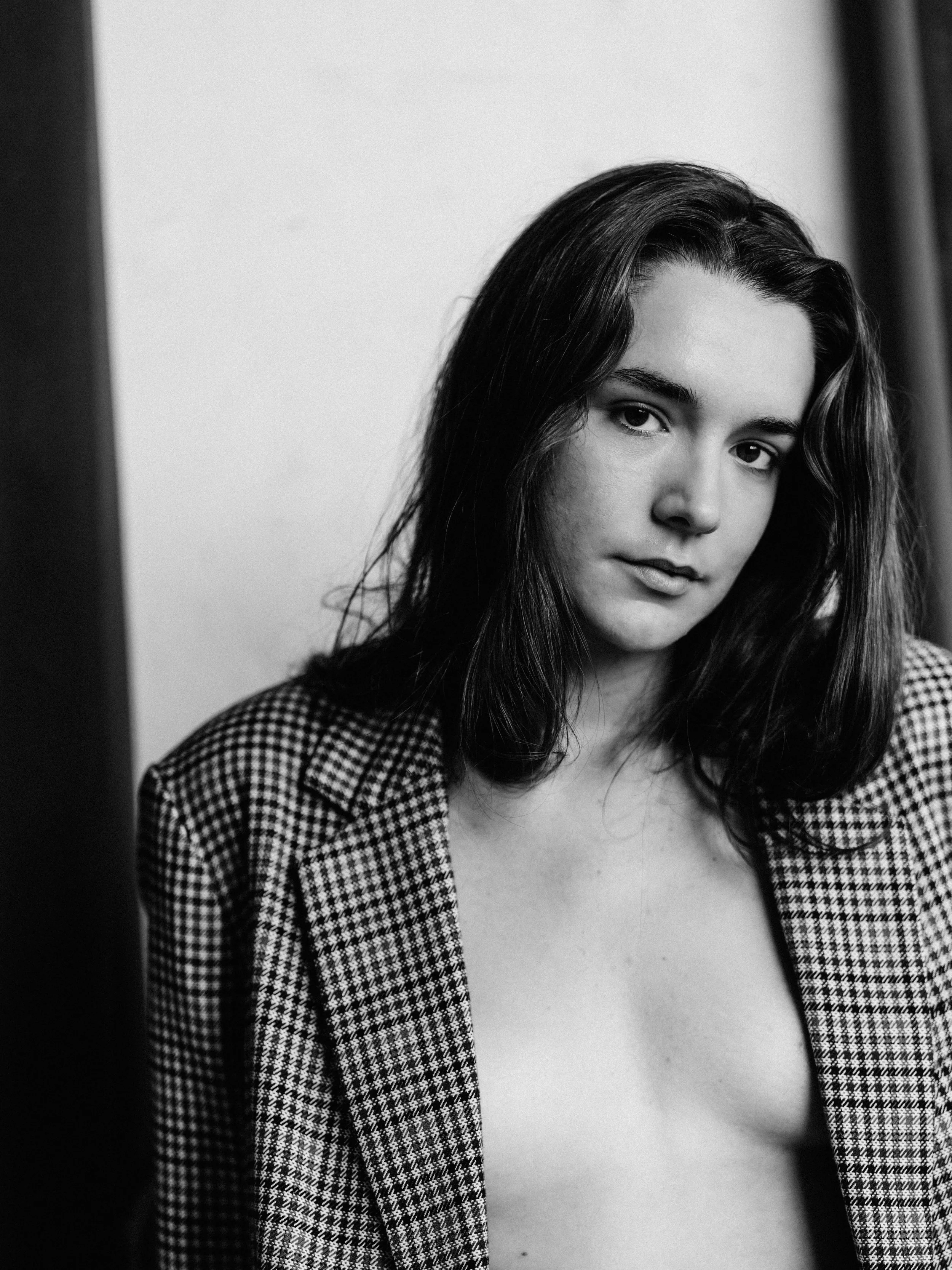 A black-and-white photograph of a woman with long, wavy hair, looking towards the camera with a neutral expression. She is wearing a houndstooth patterned blazer and a low-cut top.