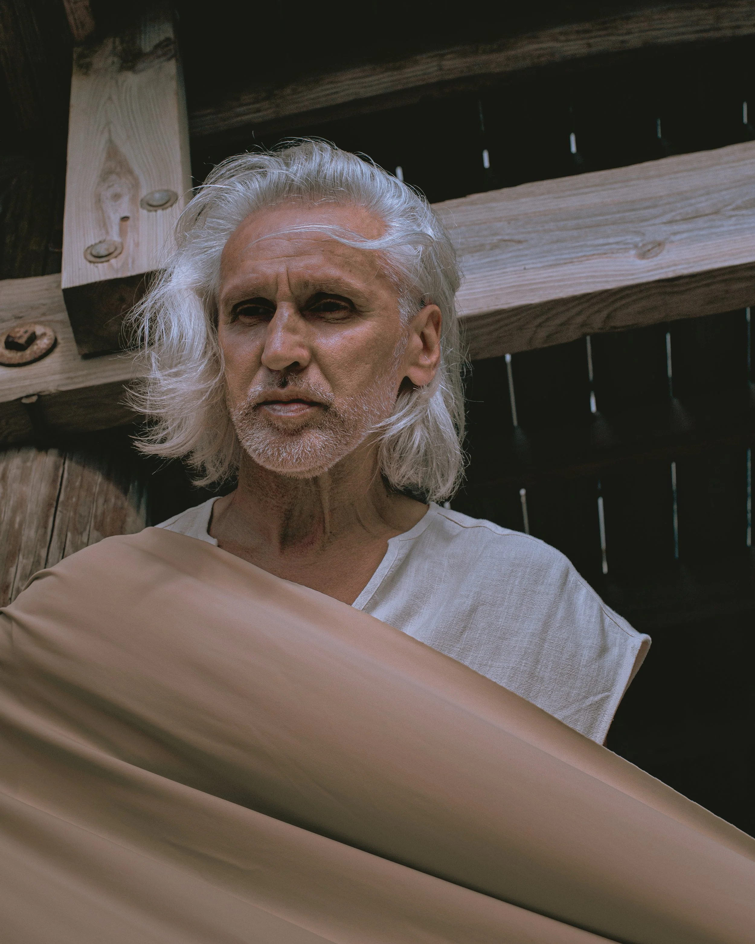 Close-up of an elderly man with long, white hair and a beard, wearing a white shirt, standing outdoors against a background of wooden beams and a black fence.
