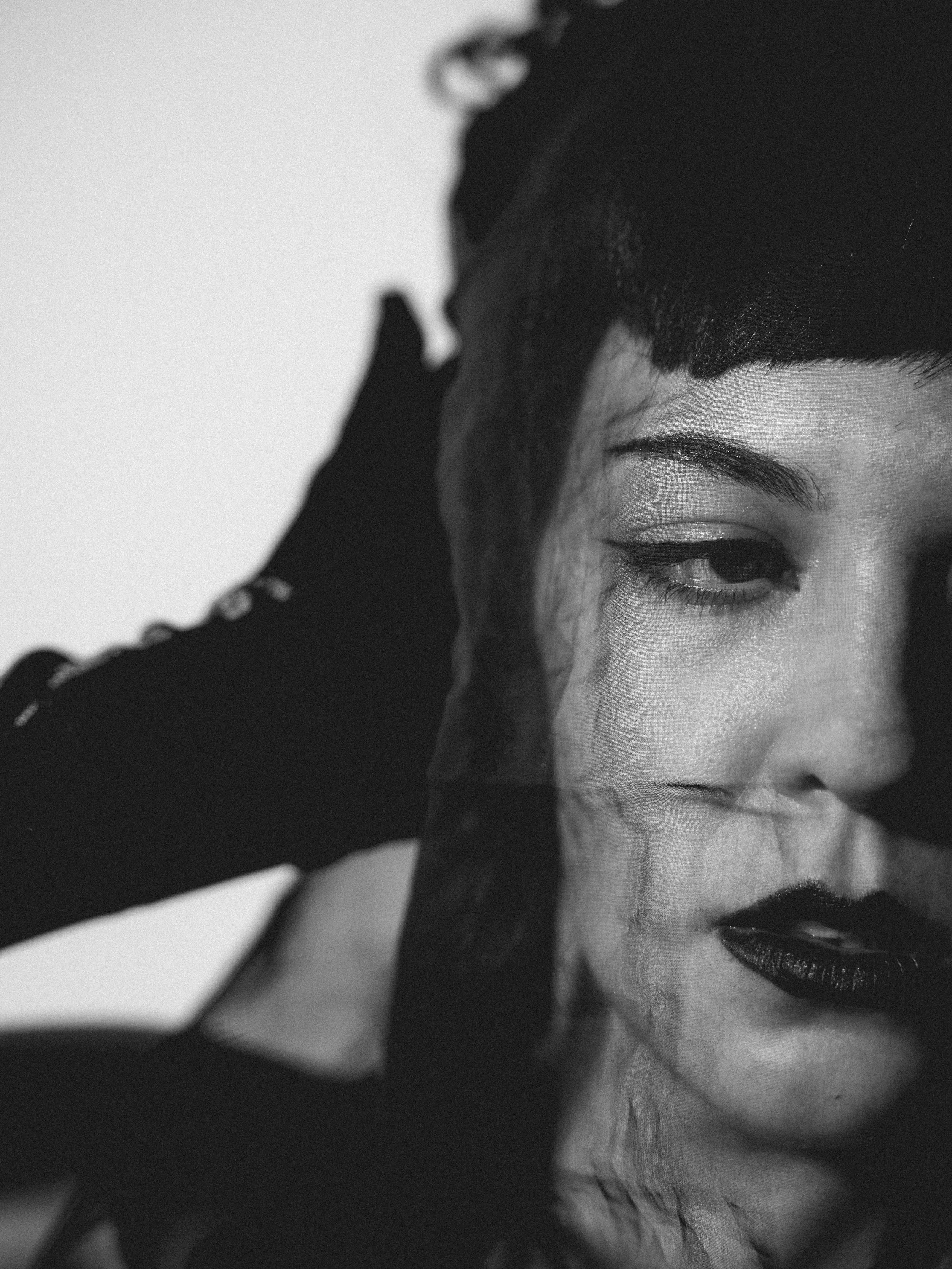 Close-up black and white photo of a woman's face, showcasing her eye with makeup, and part of her hand with a glove touching her forehead.