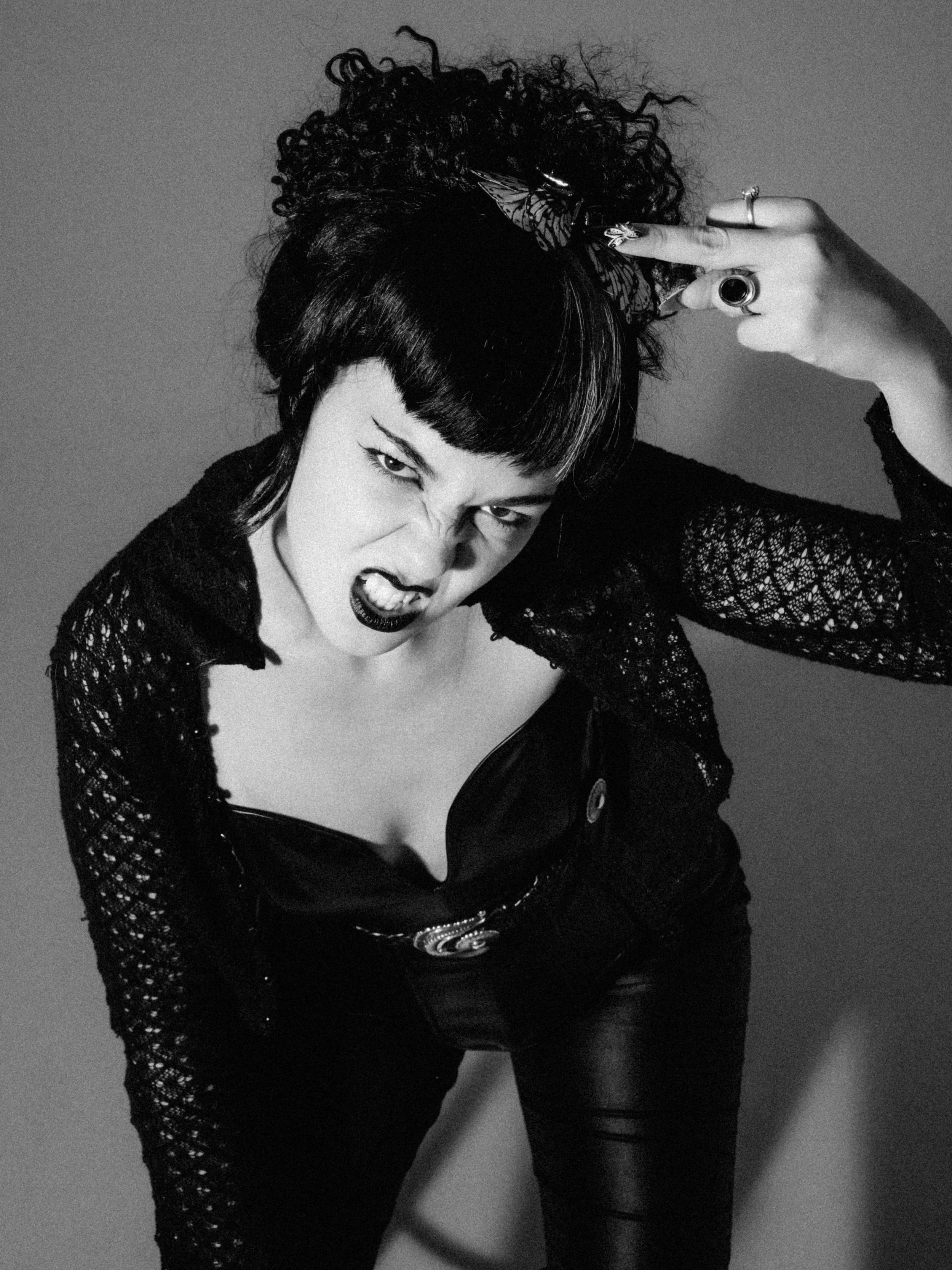 Black and white photo of a woman with curly hair in a high bun, wearing dark makeup, a lace top, and leather pants, striking a fierce pose.